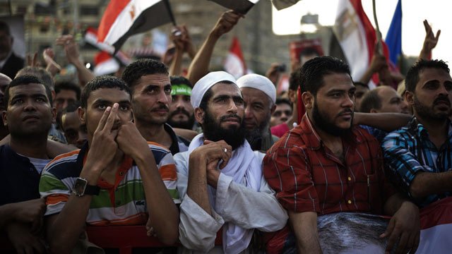L'image montre un groupe de personnes rassemblées lors d'une manifestation. On peut voir des hommes de différentes origines, certains portant des vêtements traditionnels, d'autres des t-shirts colorés. Ils semblent tous attentifs et engagés, certains levant les mains ou tenant des drapeaux. L'ambiance semble être celle d'une mobilisation ou d'une expression de soutien pour une cause. Le fond montre un environnement urbain, avec d'autres manifestants et des drapeaux visibles.
