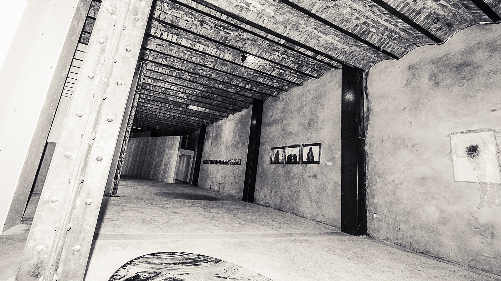 L'image présente l'intérieur d'un espace d'exposition ou d'une galerie d'art, avec de hauts plafonds en poutres de bois. Les murs sont en béton brut, créant une ambiance industrielle. Sur le mur de droite, on peut apercevoir quelques cadres accrochés, probablement contenant des œuvres d'art. Le sol est neutre, avec une texture qui semble lisse. L'éclairage met en valeur les détails architecturaux de l'intérieur, tandis que l'ensemble dégage une atmosphère contemporaine et artistique.