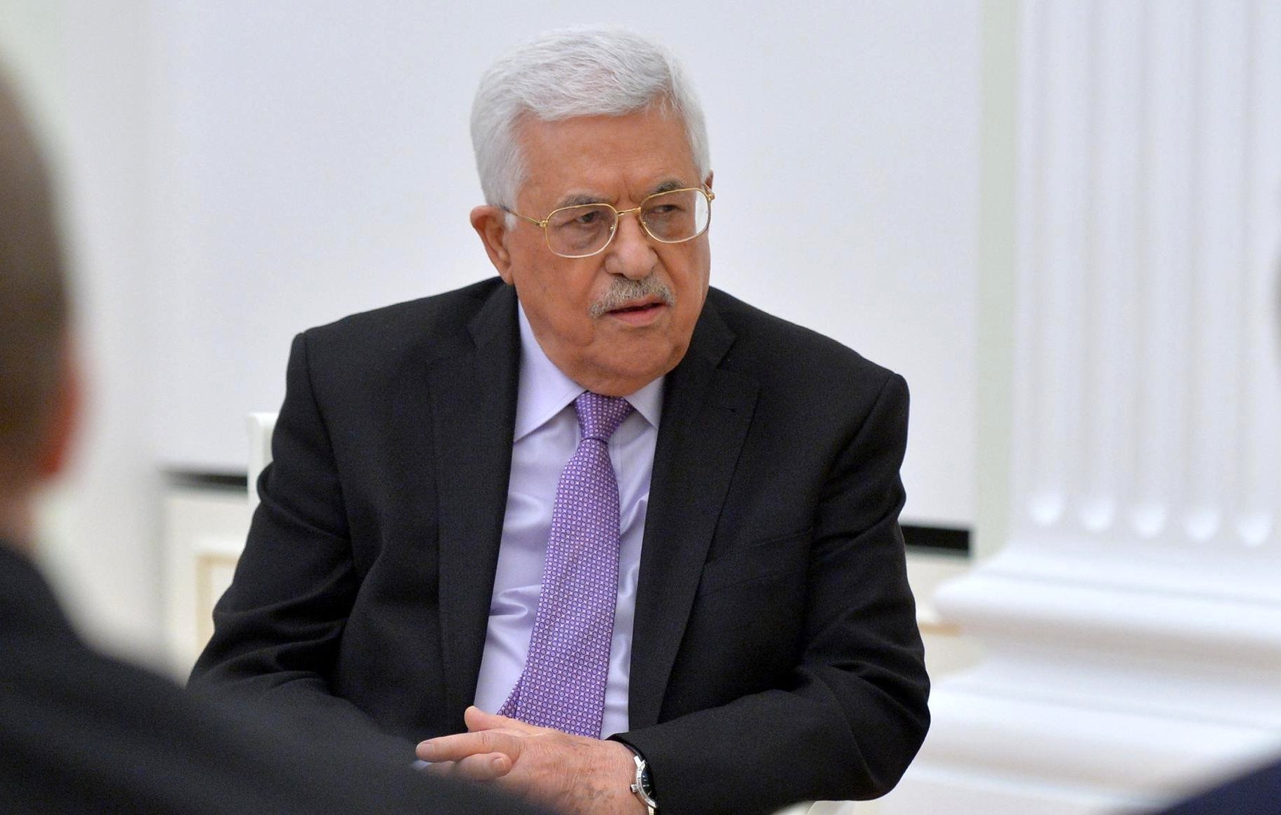 L'image montre un homme assis, portant un costume noir avec une chemise blanche et une cravate mauve. Il a des cheveux gris et porte des lunettes. L'environnement semble formel, probablement une salle de réunion. On peut noter son expression concentrée et attentif pendant la discussion.