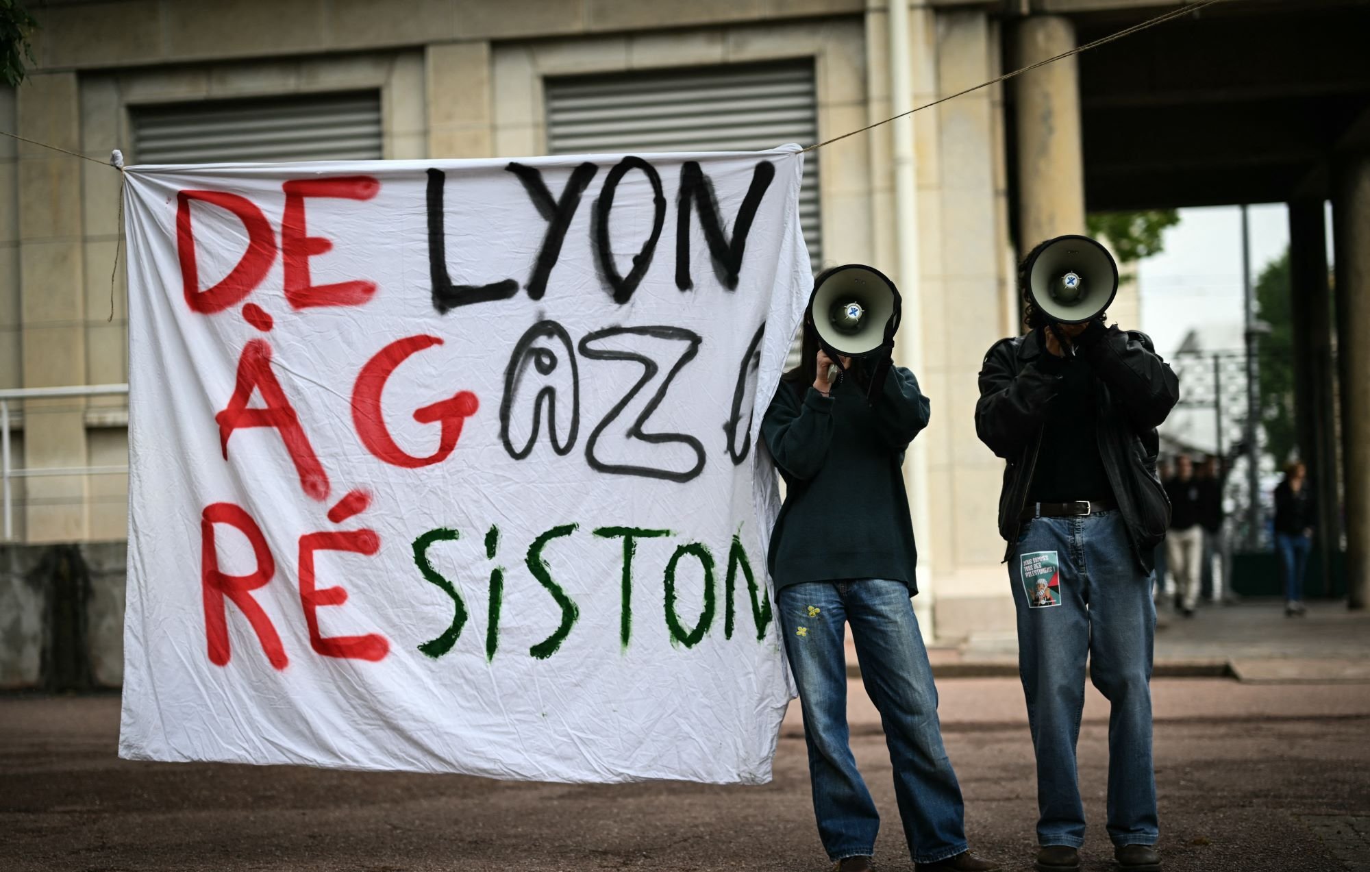 L'image montre une manifestation avec une grande pancarte accrochée qui dit "DE LYON À GAZA RÉSISTONS". Deux personnes se tiennent derrière la pancarte, chacune tenant un mégaphone. L'environnement semble urbain, et l'atmosphère suggère un engagement pour une cause.