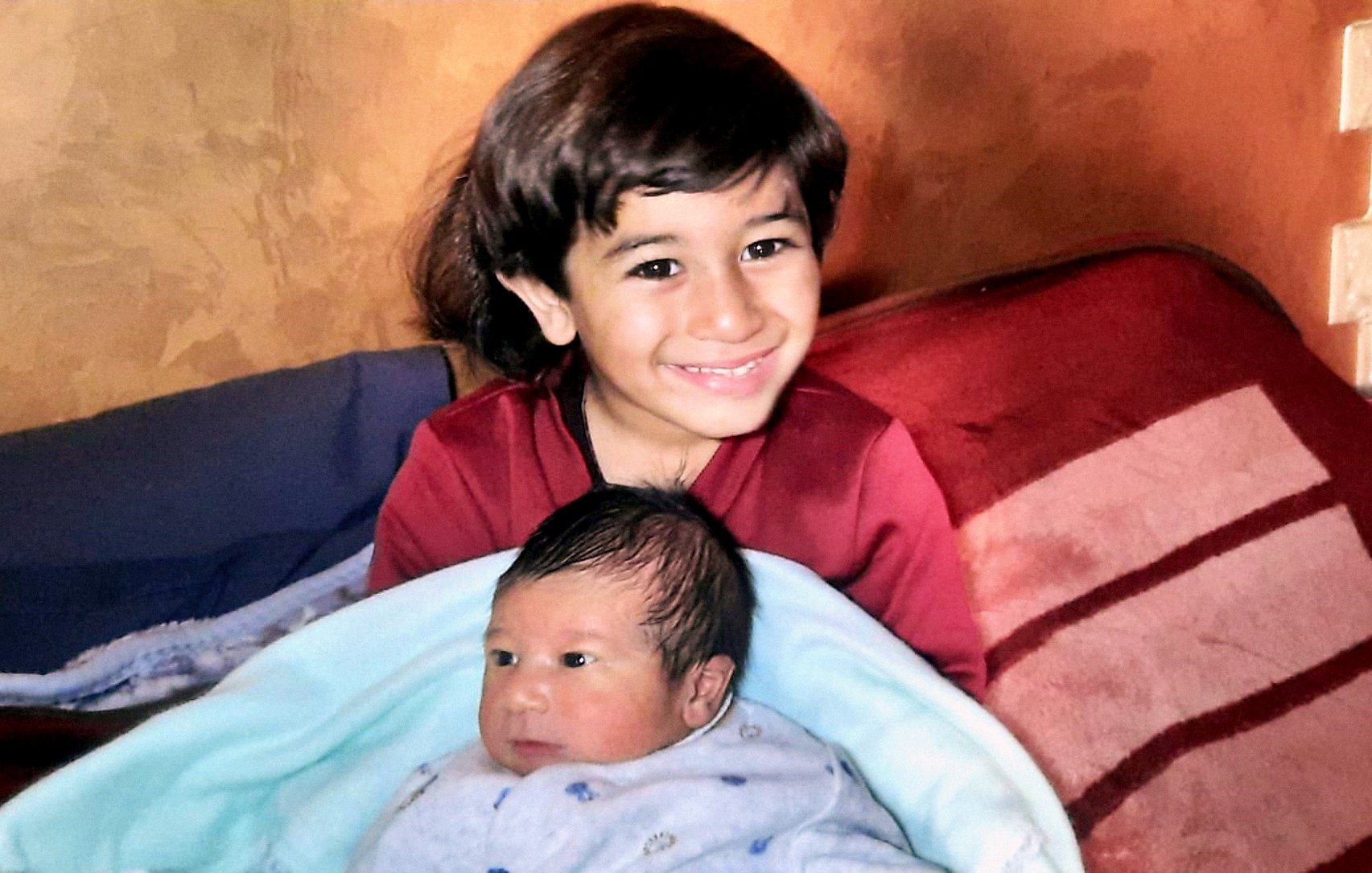 L'image montre un enfant souriant assis sur un canapé, tenant un bébé enveloppé dans une couverture bleue. L'enfant plus âgé semble heureux et fier, tandis que le bébé a une expression calme. Le décor est chaleureux avec des murs aux teintes terreuses.