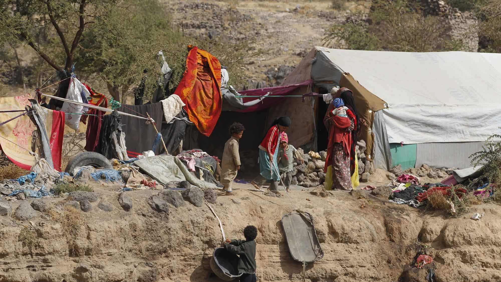 L'image montre un paysage difficile, probablement dans une région aride. On y voit des habitations précaires constituées de bâches et de morceaux de tissu, témoignant de conditions de vie modestes. Quelques enfants jouent près des abris, tandis que d'autres semblent s'affairer. L'environnement est marqué par des terrains rocheux et des éléments naturels, suggérant une situation de vulnérabilité. La scène évoque des thèmes liés à la pauvreté et à la résilience des communautés vivant dans des conditions précaires.