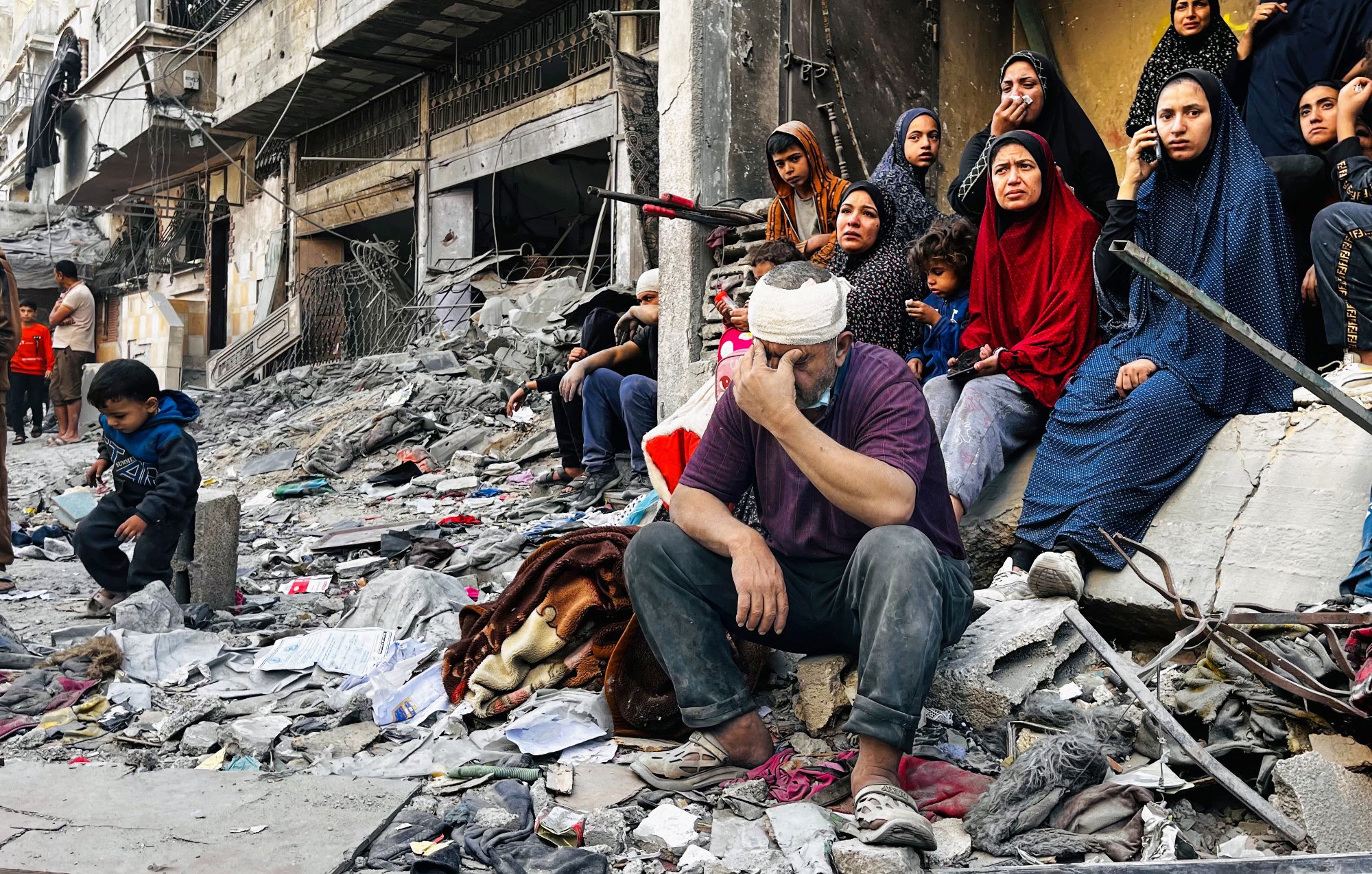 L'image montre un groupe de personnes assises sur des décombres dans une zone de conflit. Au premier plan, un homme, la tête bandée, exprime une grande tristesse ou du désespoir, tandis qu'à proximité, plusieurs femmes en vêtements sombres regardent vers l'objectif, certaines avec une expression préoccupée. Un petit garçon est assis un peu plus loin, semblant perdu dans ses pensées. L'environnement est chaotique, avec des ruines et des débris autour, suggérant des dommages importants causés par des événements récents. L'atmosphère générale est lourde, reflétant la douleur et la résilience des personnes présentes.