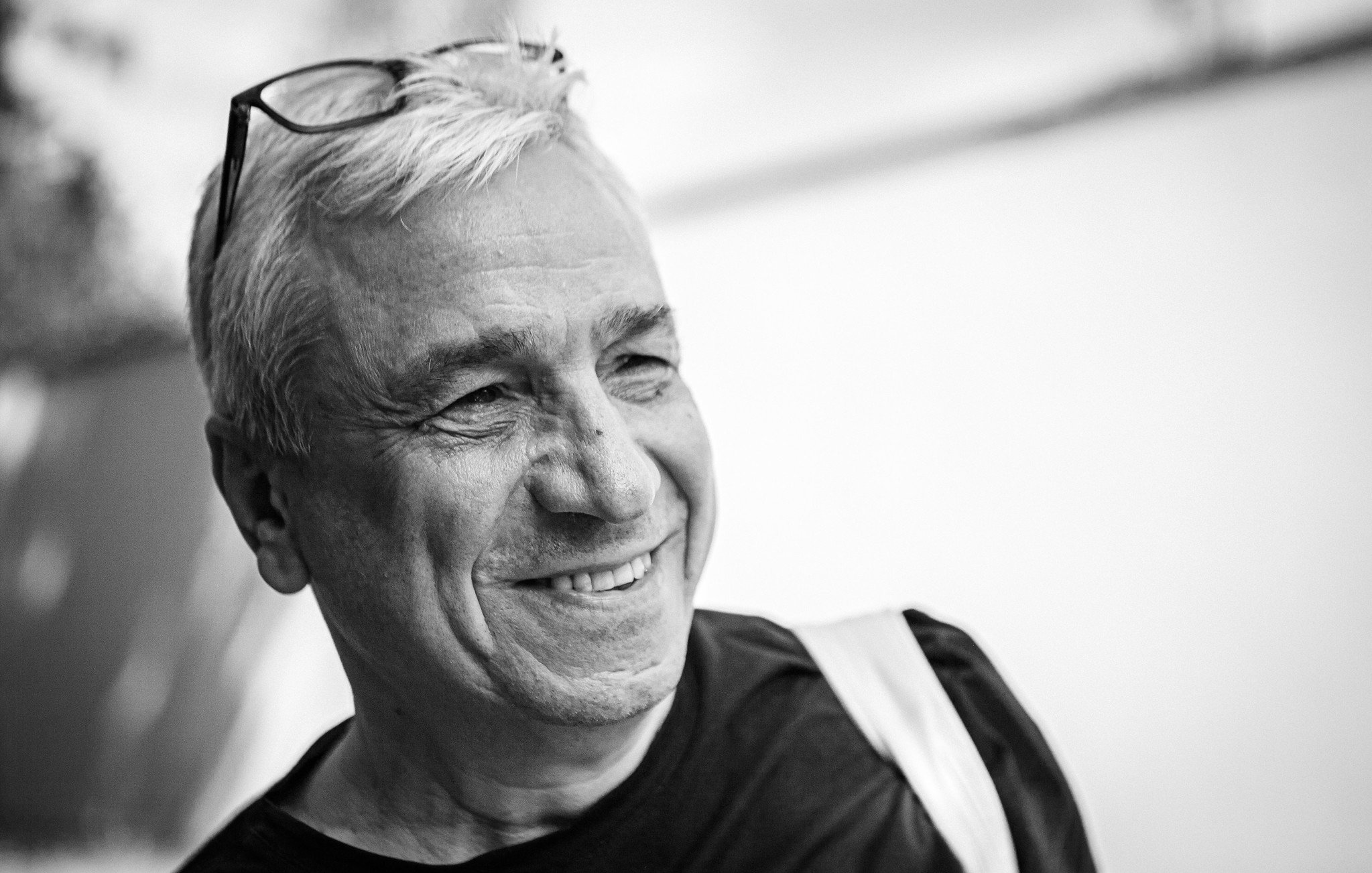 L'image montre un homme souriant. Il a des cheveux blancs et porte des lunettes sur le haut de sa tête. Son expression est joyeuse, et on peut sentir une atmosphère amicale. L'arrière-plan est flou, mettant davantage l'accent sur son visage. Il porte un t-shirt noir et semble détendu.