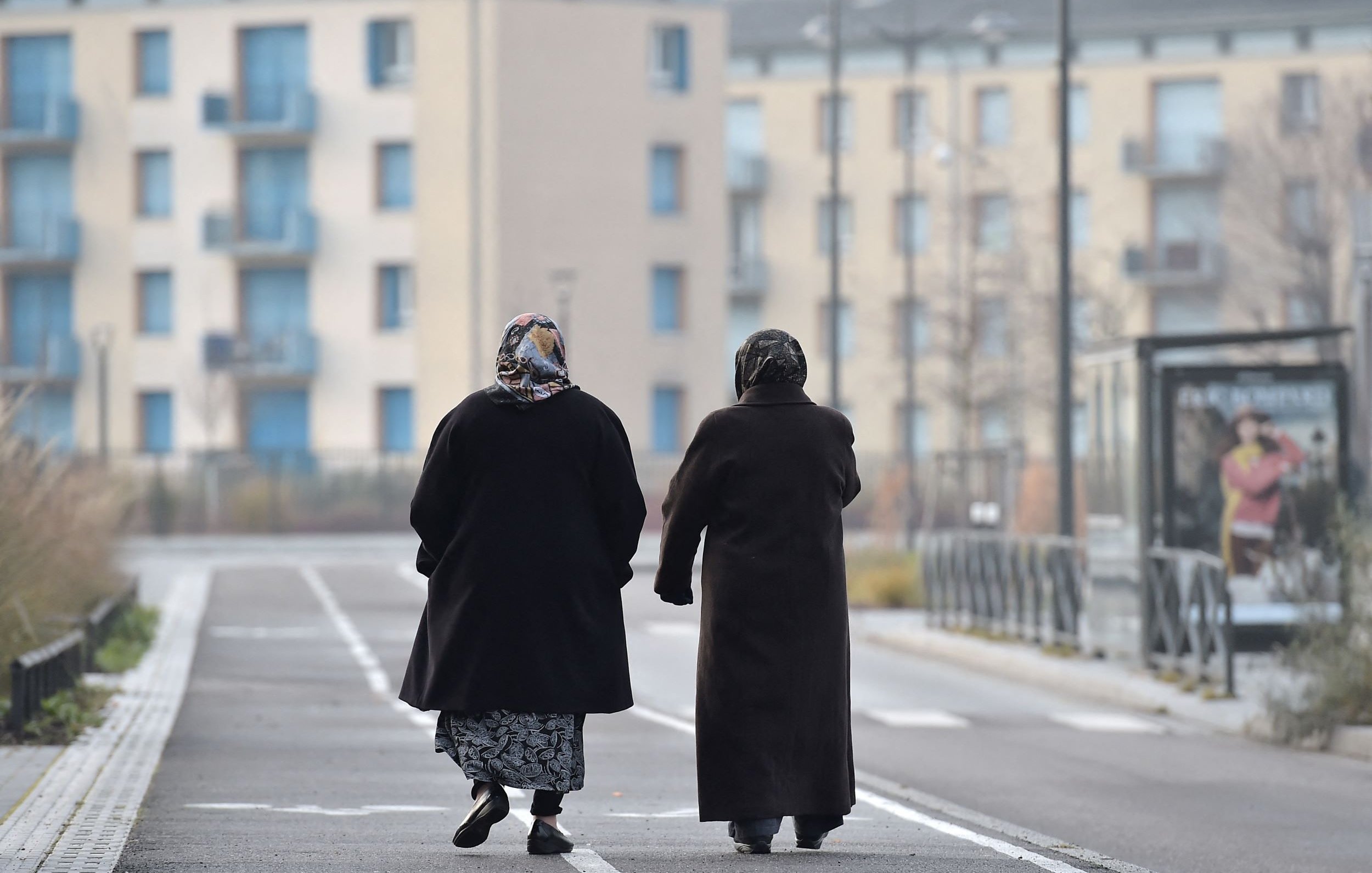 L'image montre deux femmes marchant côte à côte sur une voie urbaine. Elles portent des manteaux sombres et des foulards, ce qui suggère qu'il fait frais. En arrière-plan, on distingue des bâtiments modernes avec des balcons et des fenêtres. La scène dégage une atmosphère tranquille, avec un chemin bordé d'herbes. Leurs silhouettes semblent illustrer un moment de complicité et de camaraderie.