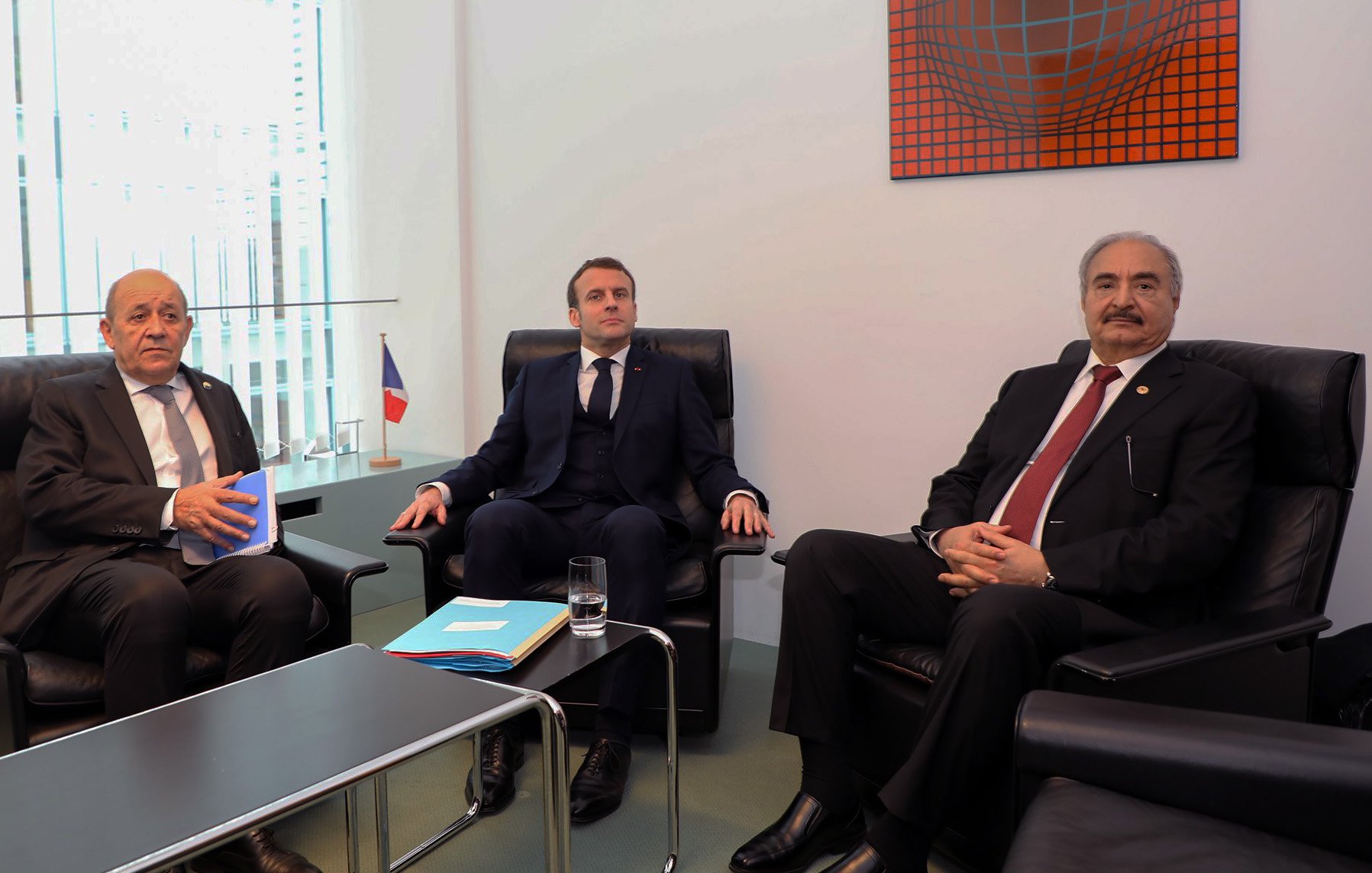 L'image montre trois hommes assis dans un cadre formel. À gauche, un homme avec des cheveux courts, habillé en costume sombre, semble tenir un document. Au centre, un homme en costume bleu regardant droit devant lui. À droite, un homme en costume noir, avec des cheveux grisonnants. Ils sont assis dans des fauteuils confortables, et il y a une table basse entre eux avec un verre d'eau. En arrière-plan, on aperçoit une œuvre d'art et un drapeau français. L'atmosphère semble sérieuse et professionnelle.