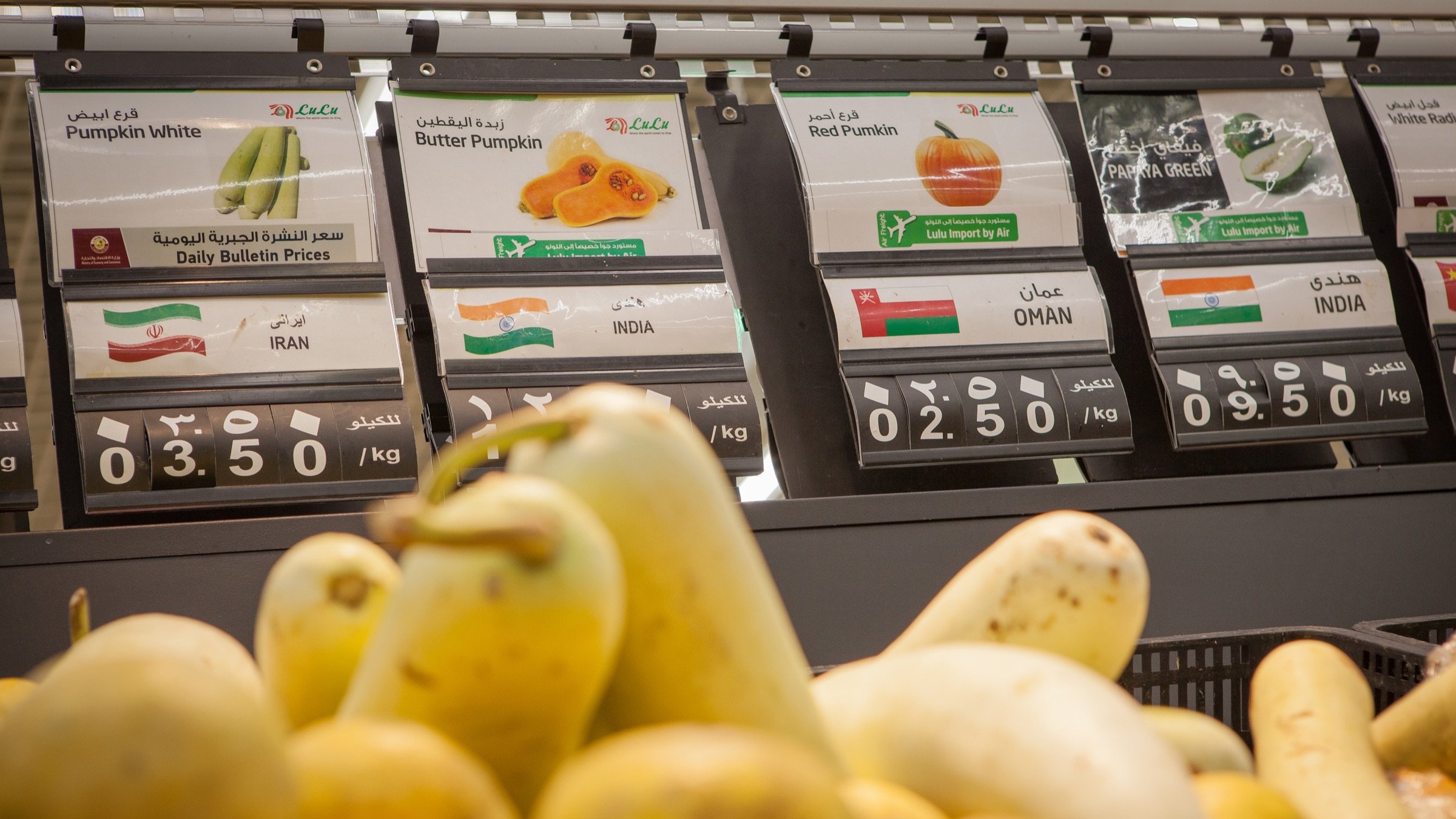 L'image présente un banc de marché dans un magasin, où divers types de courges sont exposés. Au premier plan, on voit des courges de différentes formes et couleurs. En arrière-plan, des étiquettes indiquent les prix et les origines des produits, avec des mentions comme "Courge blanche" et "Courge butternut", ainsi que des pays d'origine tels que l'Iran, Oman et l'Inde. Les prix sont affichés en kilos. L'ensemble crée une ambiance de supermarché frais et coloré.