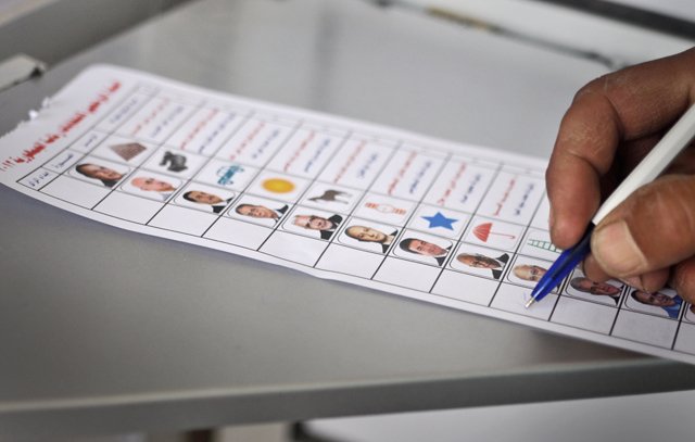 L'image montre une main tenant un stylo en train de remplir un bulletin de vote. Le bulletin affiche plusieurs colonnes, chacune contenant des photos de différentes personnes, accompagnées de symboles et de cases à cocher. Cela semble être un contexte électoral, où une personne vote pour des candidats. Le bulletin est posé sur une surface grise.