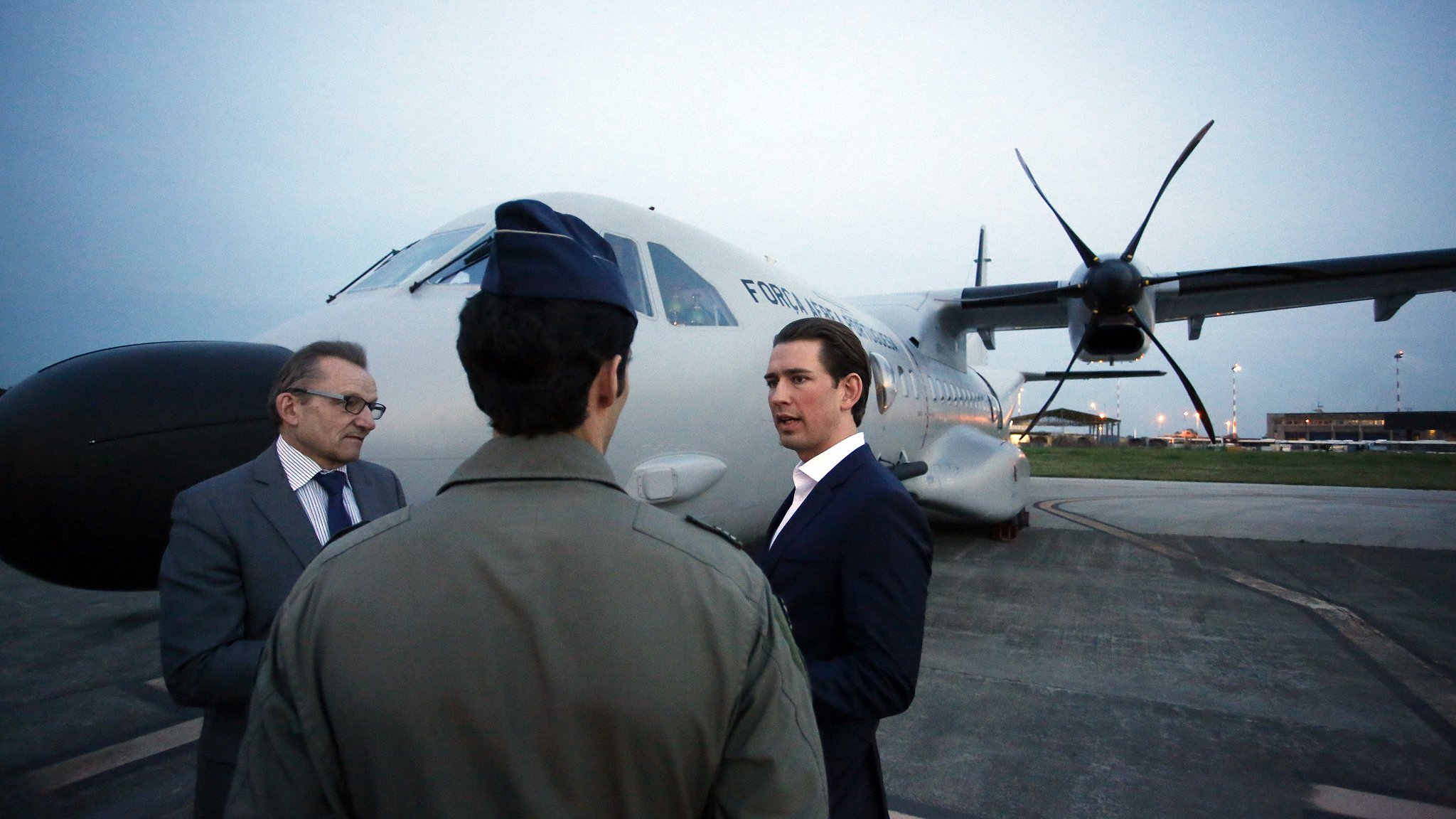 L'image montre trois hommes près d'un avion. L'un des hommes porte un uniforme militaire, tandis que les deux autres sont habillés dans des costumes. Ils semblent être en conversation, face à l'avion de transport, qui est stationné sur le tarmac à la tombée de la nuit. L'éclairage ambiant est doux, et on peut voir des lumières à l'arrière-plan, probablement d'un aéroport ou d'une base aérienne.