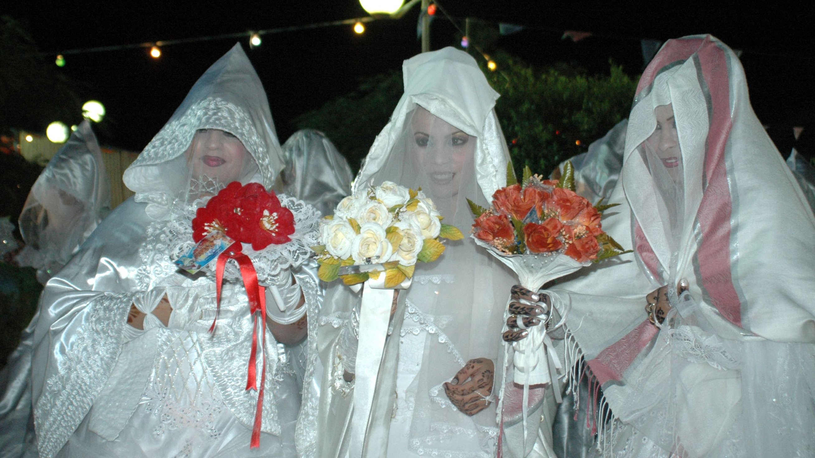 L'image montre un groupe de femmes vêtues de robes blanches et de voiles, se tenant ensemble dans une ambiance nocturne. Elles tiennent chacune des bouquets de fleurs, composés de roses et d'autres fleurs colorées. L'éclairage doux crée une atmosphère festive, et des guirlandes lumineuses sont visibles en arrière-plan. Leurs visages sont en partie cachés par les voiles, ajoutant une aura de mystère à la scène.