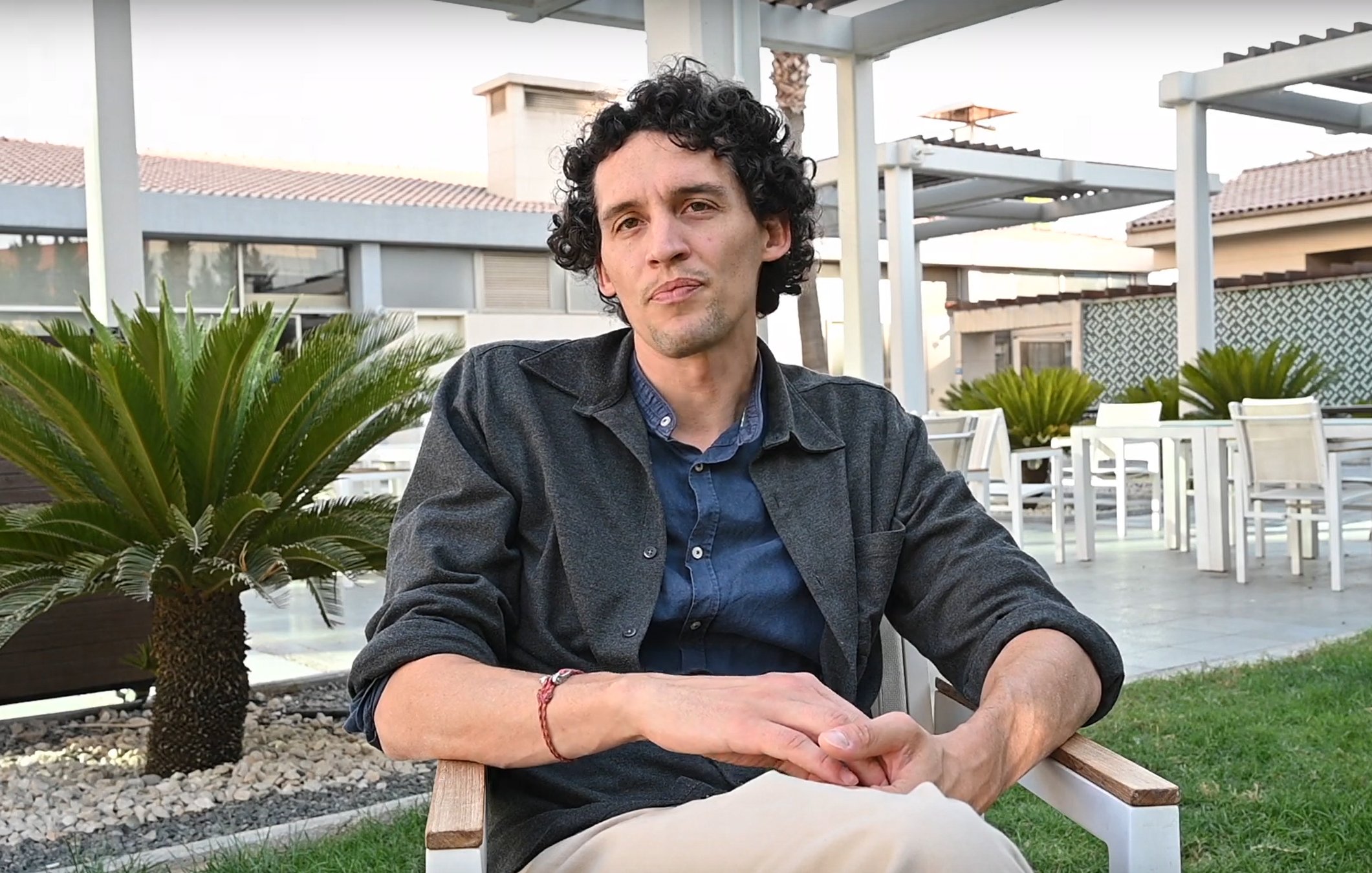 L'image montre un homme assis sur une chaise en bois dans un environnement extérieur. Il a des cheveux bouclés et porte une chemise sombre sur un t-shirt. Il est assis dans un cadre paisible, avec des plantes vertes autour de lui et une terrasse visible en arrière-plan. L'ambiance semble détendue et agréable.