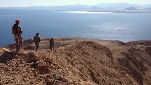 L'image montre un paysage désertique avec des militaires en patrouille sur une crête surplombant une étendue d'eau. Le terrain est aride et rocheux, avec des montagnes en arrière-plan. Les soldats portent des uniformes et des casques, et semblent observer l'horizon, avec un ciel clair et ensoleillé qui ajoute à l'atmosphère de la scène.
