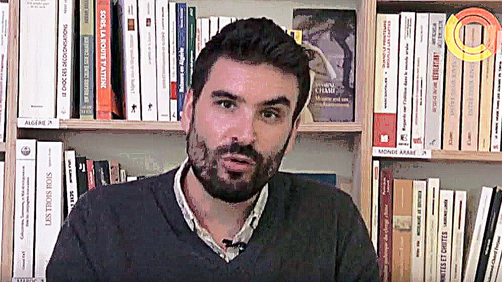 L'image montre un homme assis devant une étagère remplie de livres. Il a une barbe et porte un pull gris. L'expression de son visage semble sérieuse et il semble s'adresser à un auditoire ou à une caméra. L'arrière-plan est constitué de nombreux livres, suggérant un environnement intellectuel ou académique.