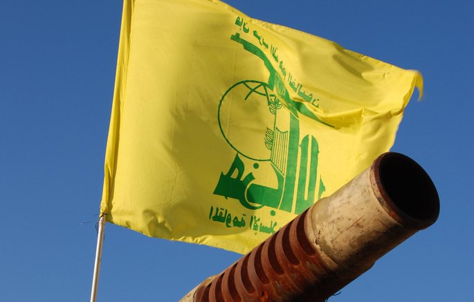 L'image montre un canon pointant vers le ciel, avec un drapeau jaune flottant au-dessus. Sur le drapeau, il y a des inscriptions en arabe ainsi qu'un logo symbolique. Le fond est bleu, suggérant une journée ensoleillée. Le canon présente des signes d'usure, avec une surface rouillée.
