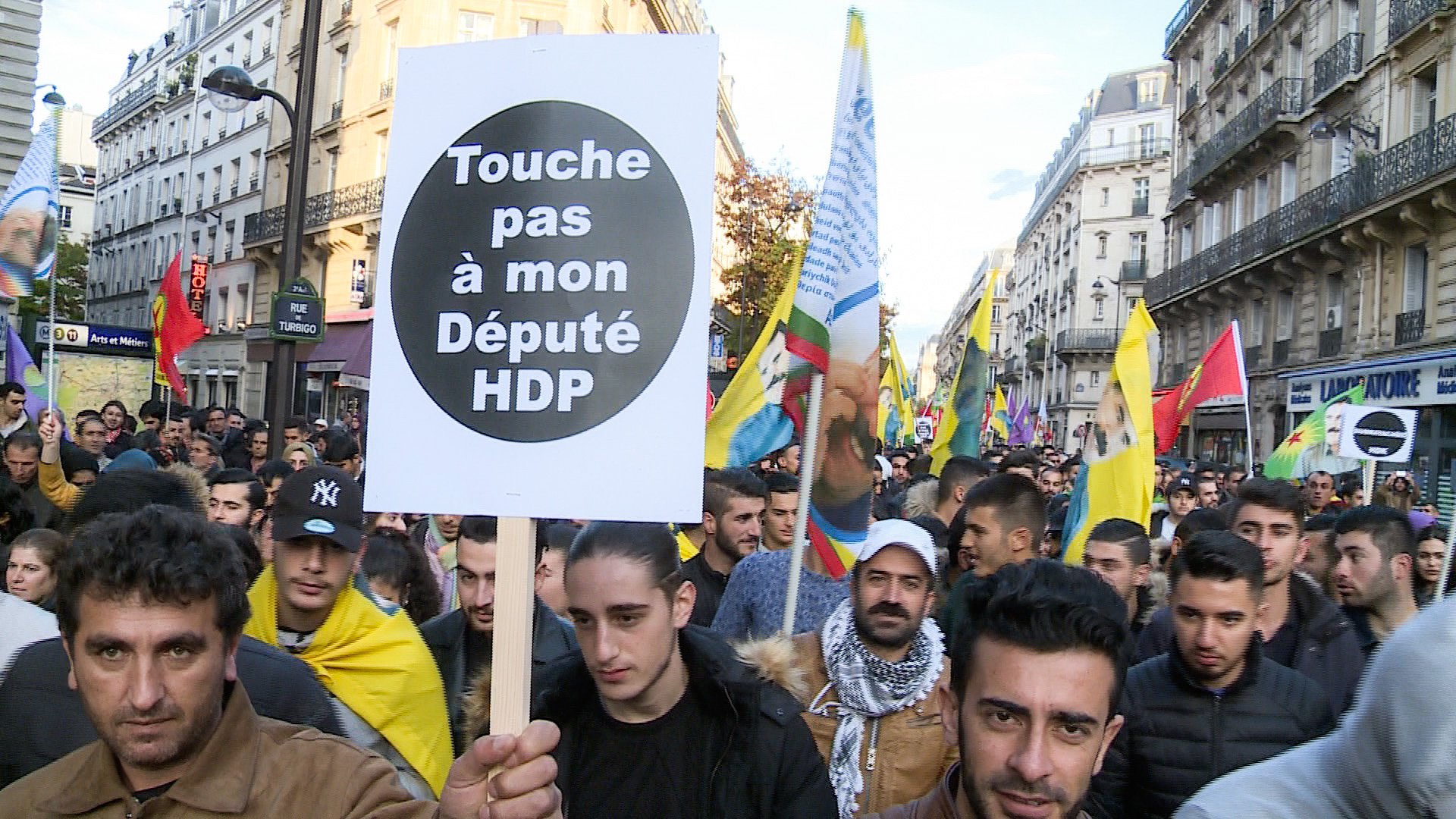 L'image montre une manifestation de foule dans une rue, où des personnes tiennent des pancartes et des drapeaux. Au premier plan, un manifestant porte une grande pancarte avec le message "Touche pas à mon Député HDP". L'ambiance semble être celle d'une protestation politique, impliquant probablement des questions liées au parti HDP (Parti démocratique des peuples) en Turquie. Les participants semblent engagés et déterminés.