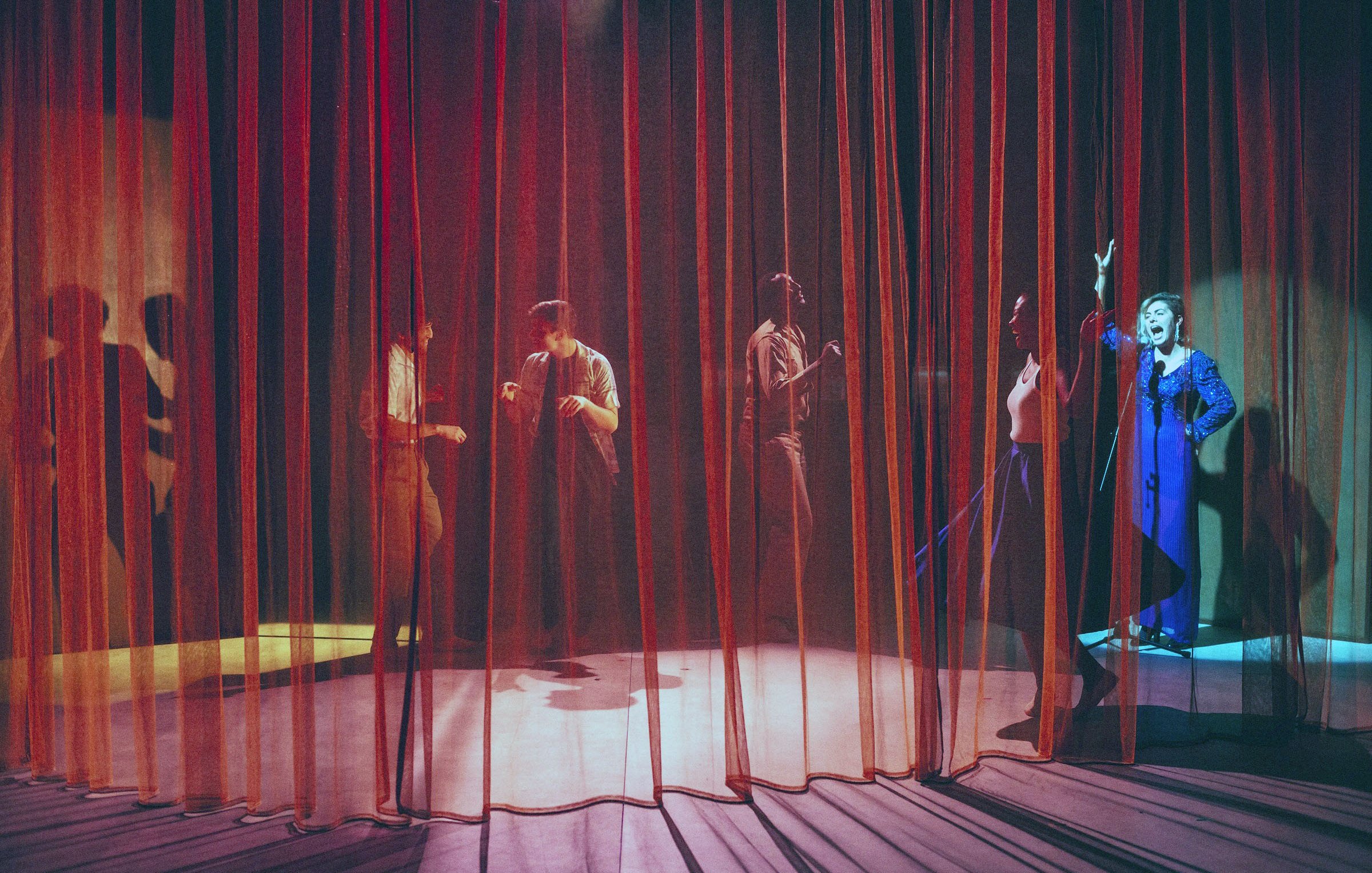 L'image montre une scène de théâtre avec des rideaux en velours rouge et orange qui créent une atmosphère intimiste. On aperçoit plusieurs silhouettes de personnes derrière les rideaux, engagées dans des activités qui semblent dynamiques et expressives. L'un des personnages, visible à droite, porte un costume bleu et semble gesticuler avec enthousiasme. Les jeux de lumière entre les rideaux ajoutent une ambiance dramatique à la scène, accentuant les mouvements des acteurs.