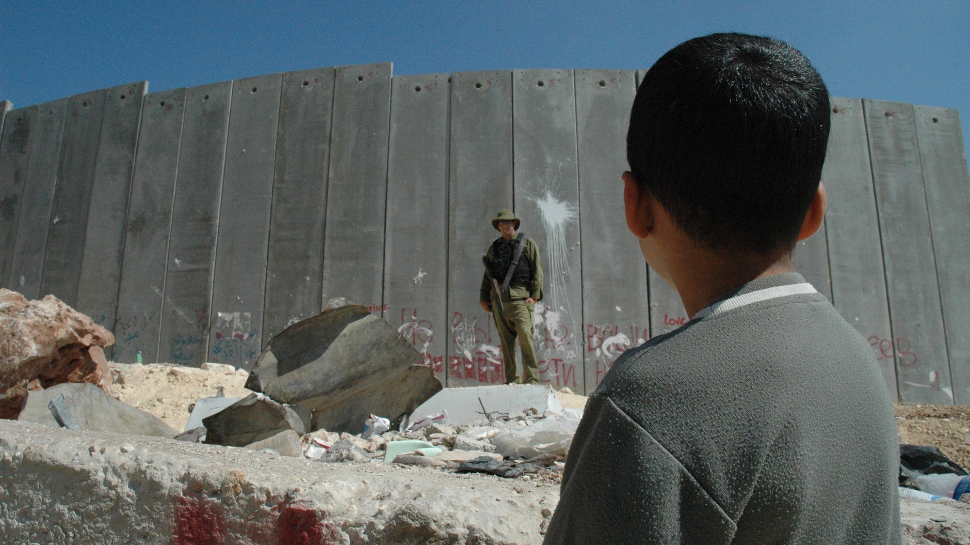 L'image montre un jeune garçon observant un soldat qui se tient devant un mur en béton. Le mur est marqué par des graffitis, suggérant un environnement de conflit ou de tension. Des débris sont dispersés au sol, et le ciel est dégagé, ce qui contraste avec la gravité de la scène. Le regard du garçon semble interrogateur ou curieux face à la situation qui l'entoure.