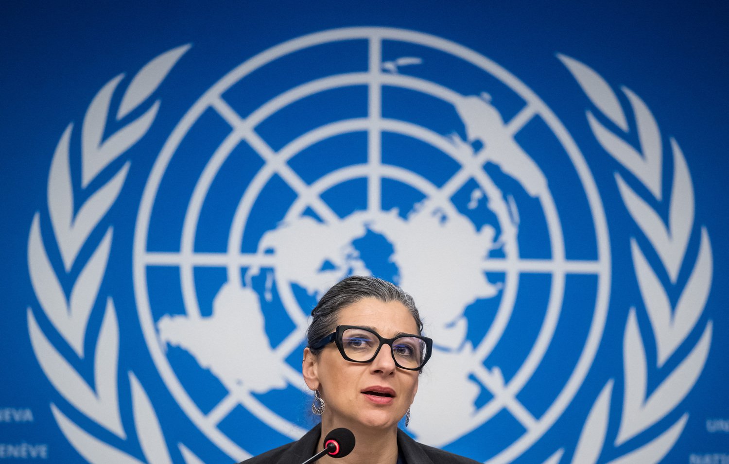 L'image montre une femme parlant devant un microphone, avec un fond représentant le logo des Nations Unies. Elle porte des lunettes et a les cheveux attachés. Son expression semble concentrée, et elle est probablement en train de faire une déclaration ou de répondre à des questions. Le décor bleu est typique des conférences de presse internationales.