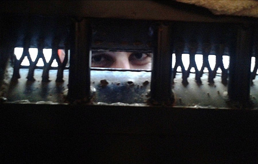 L'image montre un regard émergeant à travers une grille ou une ouverture sombre. Les yeux de la personne sont bien visibles, tandis que le reste de son visage est caché par l'ombre ou la structure. Les bords de l'ouverture semblent usés, ajoutant une atmosphère de confinement ou de mystère à la scène.