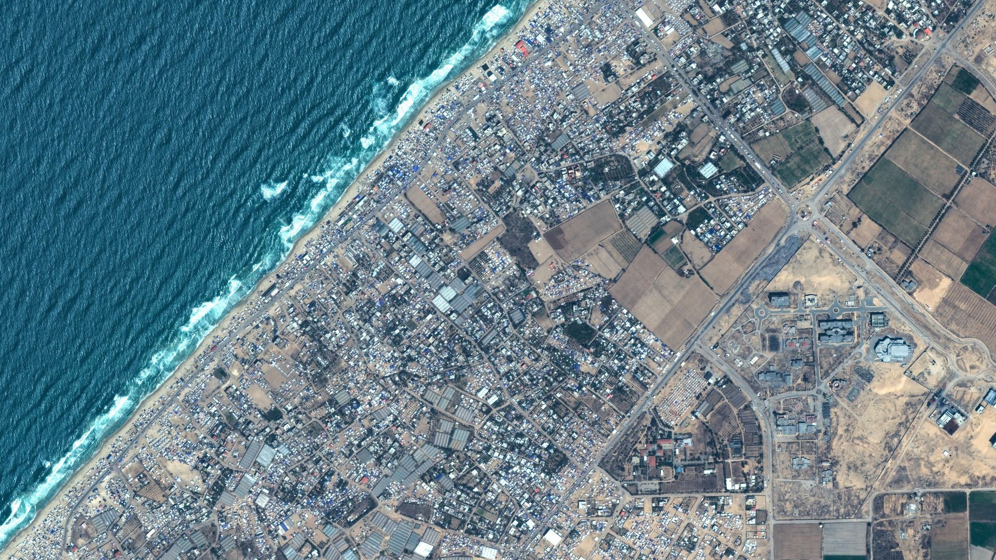 L'image montre une vue aérienne d'une zone côtière. En haut à gauche, on peut voir l'océan avec des vagues qui se brisent sur la plage. La partie inférieure de l'image présente des terrains variés, incluant des espaces urbanisés avec des routes et des bâtiments, ainsi que des zones agricoles avec des champs. La transition entre la zone bâtie et la nature est également visible. C'est une représentation d'un paysage où les zones urbaines rencontrent la côte.