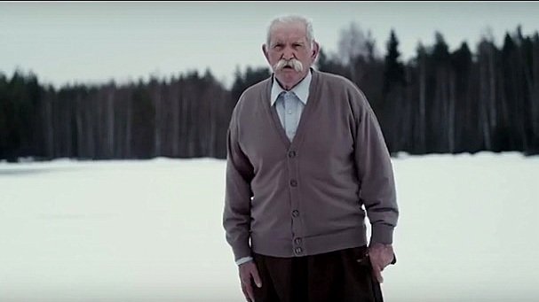 L'image montre un homme âgé se tenant debout sur un terrain enneigé. Il porte un pull en cardigan gris et un pantalon, et il a une moustache bien fournie. En arrière-plan, on peut voir une forêt de conifères qui ajoute une ambiance tranquille et froide à la scène. L'homme semble contemplatif, regardant droit devant lui dans un paysage hivernal.