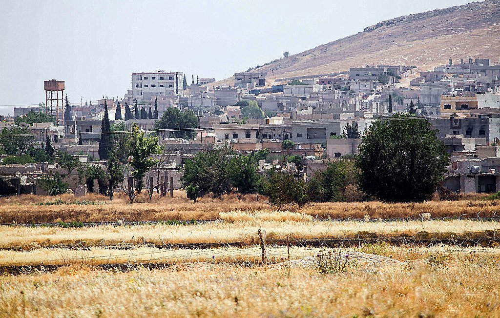L'image montre un paysage rural avec des bâtiments en arrière-plan, probablement une petite ville ou un village. Au premier plan, il y a des champs avec de l'herbe sèche ou des cultures en attente de récolte. On peut voir une colline à l'arrière-plan, ainsi qu'un réservoir d'eau. La végétation est présente, avec quelques arbres éparpillés. L'atmosphère semble calme et ensoleillée, dégageant une ambiance de milieu rural.