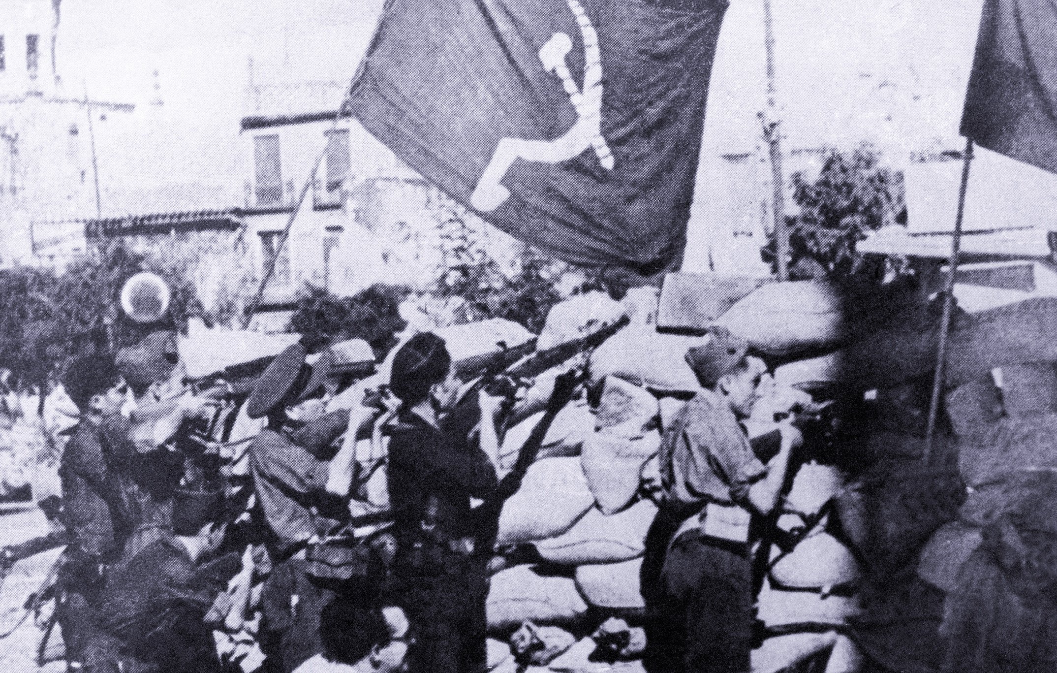 Cette image en noir et blanc montre un groupe de soldats ou de miliciens en position de tir, probablement lors d'un conflit. Ils se trouvent derrière un mur de sacs de sable, tout en tenant des fusils. En arrière-plan, des drapeaux avec un symbole communiste flottent, ce qui suggère un contexte politique et historique marqué par des idéologies de gauche. L'ambiance de la scène semble intense et sérieuse, indiquant un moment de bataille ou de lutte.