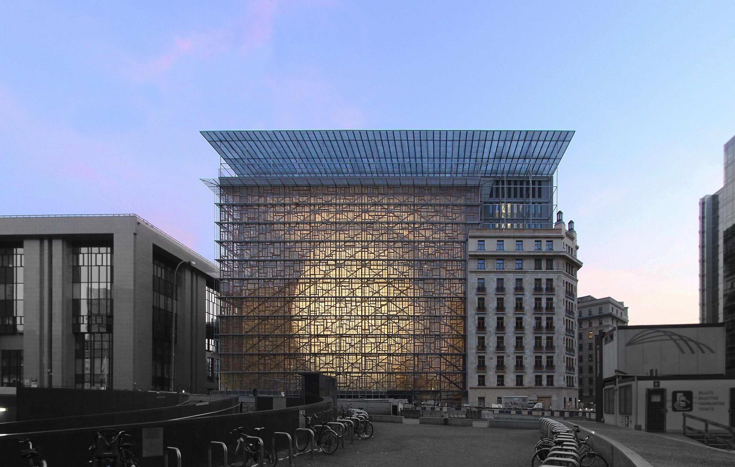 L'image montre un bâtiment moderne avec une structure en verre et en métal, se dressant au centre d'un environnement urbain. La façade réfléchissante crée un effet de transparence et de lumière, tandis que le ciel présente des nuances de bleu et de rose, suggérant un moment au crépuscule. Au premier plan, on aperçoit des vélos alignés, ajoutant une touche de vie à la scène. À gauche, il y a un autre bâtiment avec un style architectural plus traditionnel, contrastant avec la modernité du bâtiment principal.