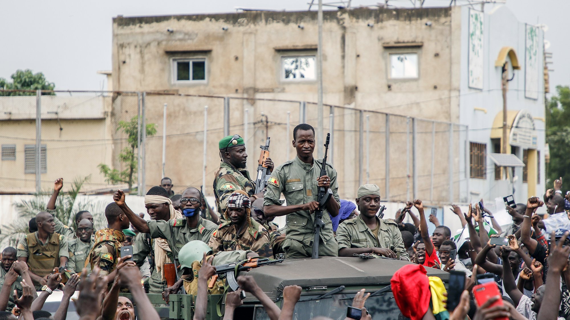 L'image montre un groupe de soldats, armés, sur un véhicule militaire, entourés par une foule enthousiaste. Les soldats semblent célébrer ou soutenir une cause, tandis que les membres de la foule lèvent les bras et expriment leur joie ou leur enthousiasme. En arrière-plan, on peut apercevoir des bâtiments et une atmosphère de mobilisation populaire.