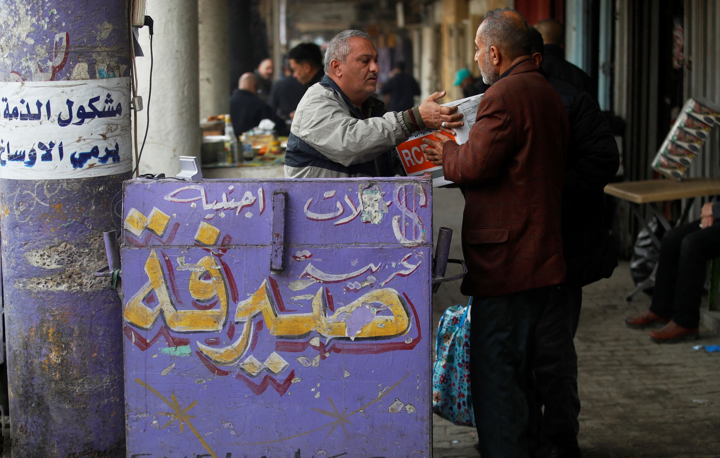 L'image montre un commerce dans une rue animée. On y voit un homme en train de discuter avec un autre, probablement en train d'échanger de l'argent ou des informations. En arrière-plan, des personnes sont assises à des tables, et on peut voir des étals remplis de marchandises. Le mur est décoré de graffitis et d'inscriptions en arabe, ainsi qu'une mention en anglais "EXCHANGE", ce qui indique probablement qu'il s'agit d'un bureau de change. L'ambiance semble vivante et typique d'un marché ou d'un bazar.