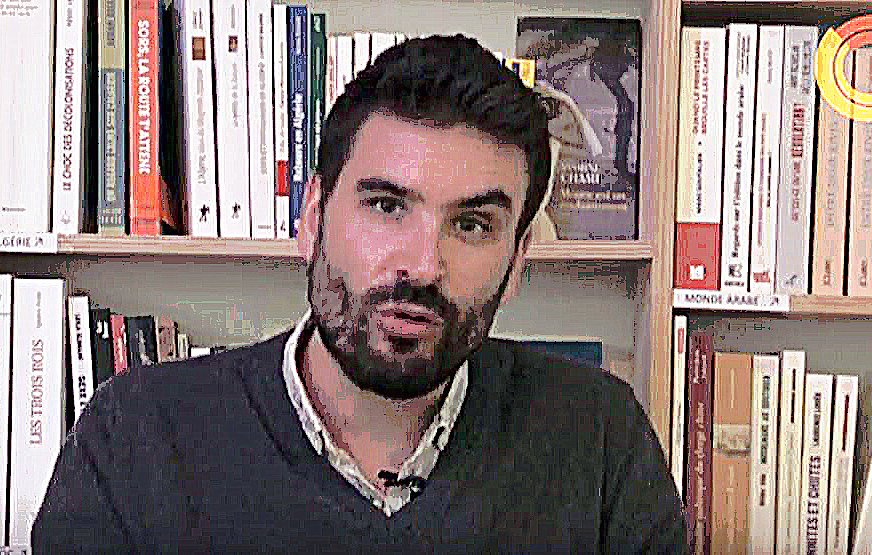 L'image montre un homme assis devant une étagère remplie de livres. Il a une barbe et porte un pull gris. L'expression de son visage semble sérieuse et il semble s'adresser à un auditoire ou à une caméra. L'arrière-plan est constitué de nombreux livres, suggérant un environnement intellectuel ou académique.