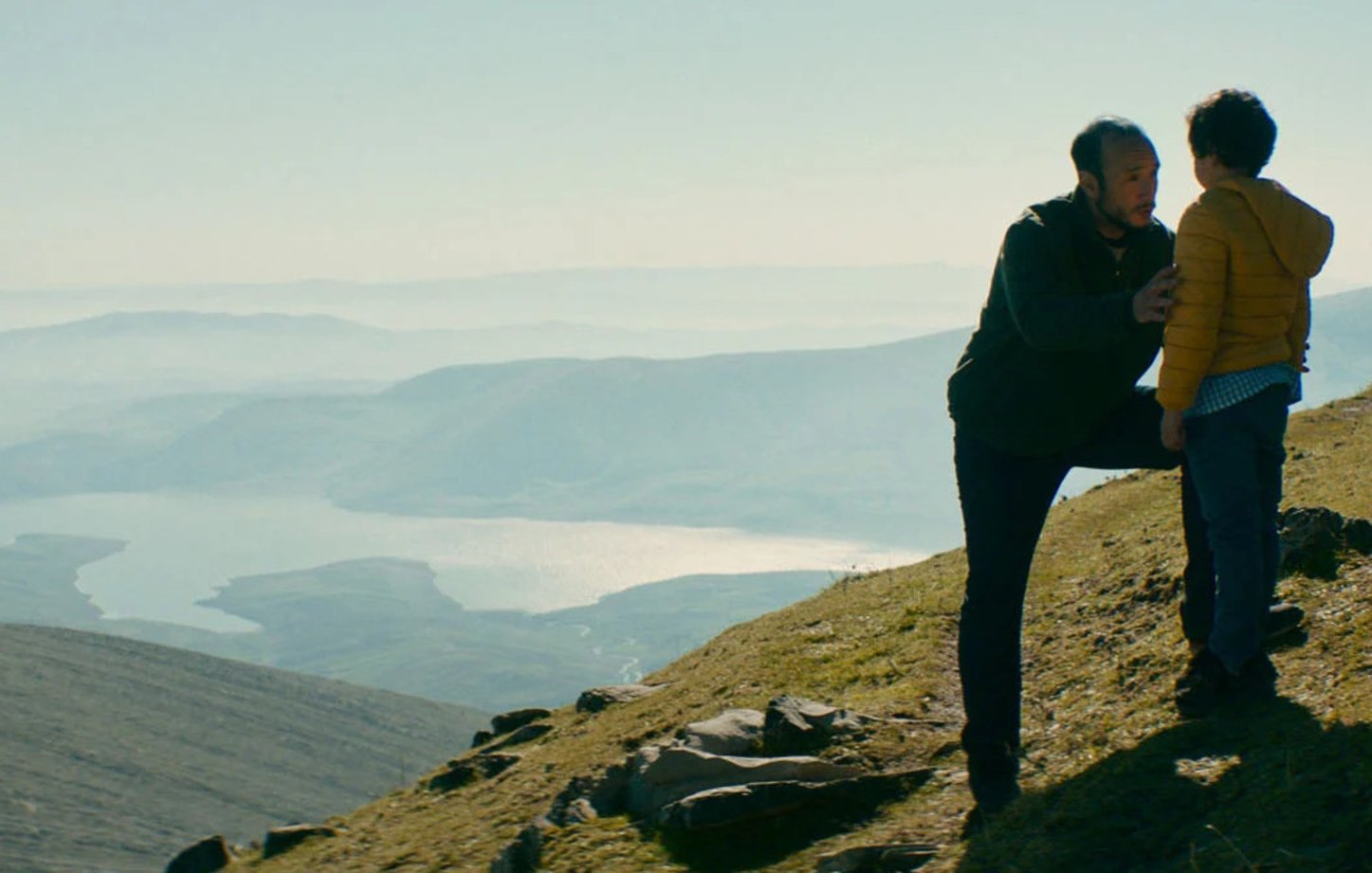 L'image montre un homme et un enfant sur une colline, face à un paysage naturel vaste et montagneux. L'homme semble parler à l'enfant, peut-être pour lui donner des conseils ou partager un moment d'intimité. En arrière-plan, on aperçoit un lac scintillant et des montagnes, le tout baigné par une lumière douce, ce qui crée une atmosphère sereine et contemplative. La scène évoque des thèmes de connexion et de découverte en pleine nature.