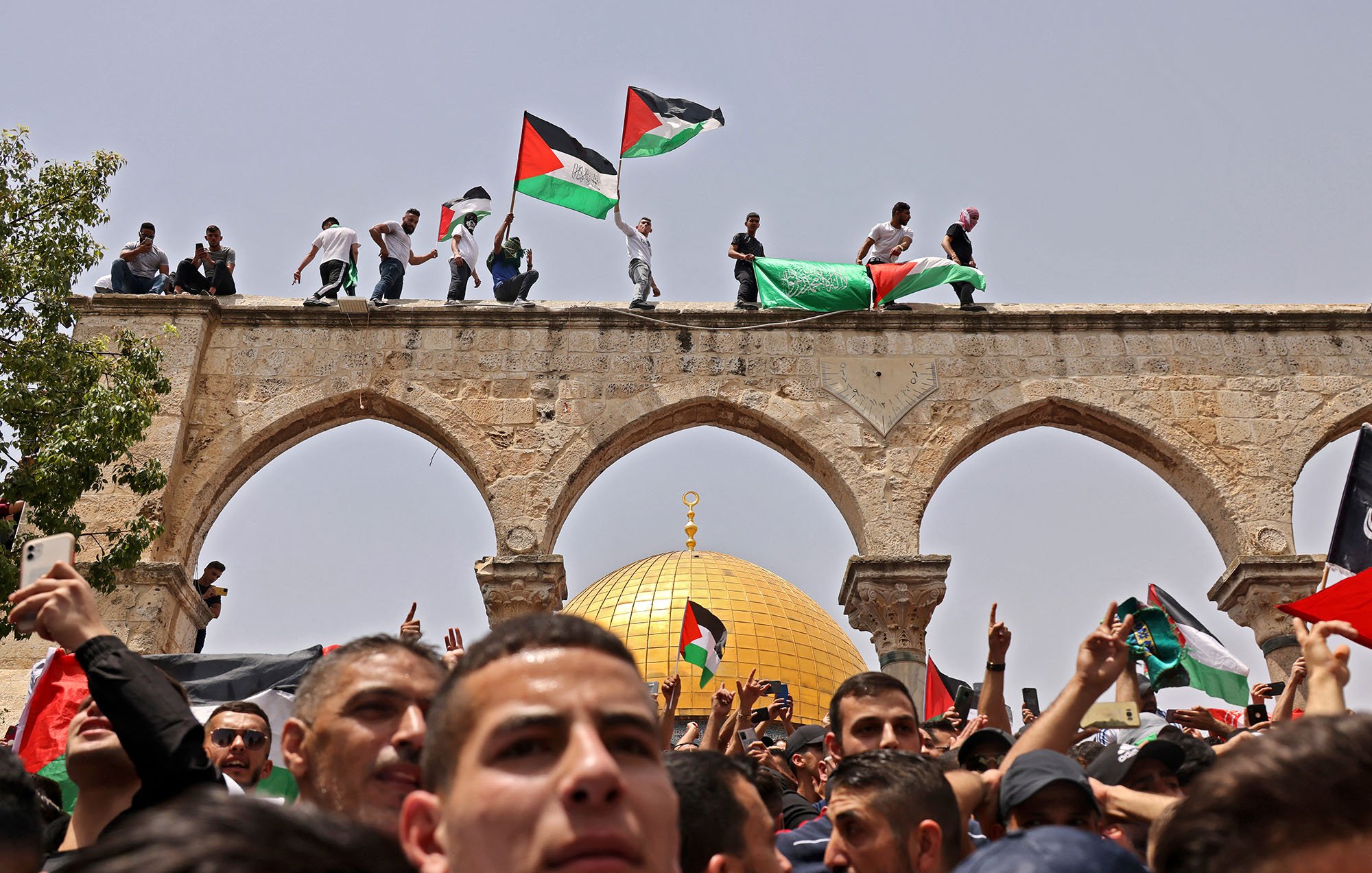 L'image montre une scène de rassemblement animé, avec un groupe de personnes portant des drapeaux palestiniens. Certaines personnes sont perchées sur un arc, tandis que la foule se trouve en dessous. En arrière-plan, on peut voir un dôme doré, probablement le Dôme du Rocher, un site emblématique de Jérusalem. L'atmosphère semble être celle d'une manifestation ou d'une célébration, avec une forte présence de symboles nationaux.