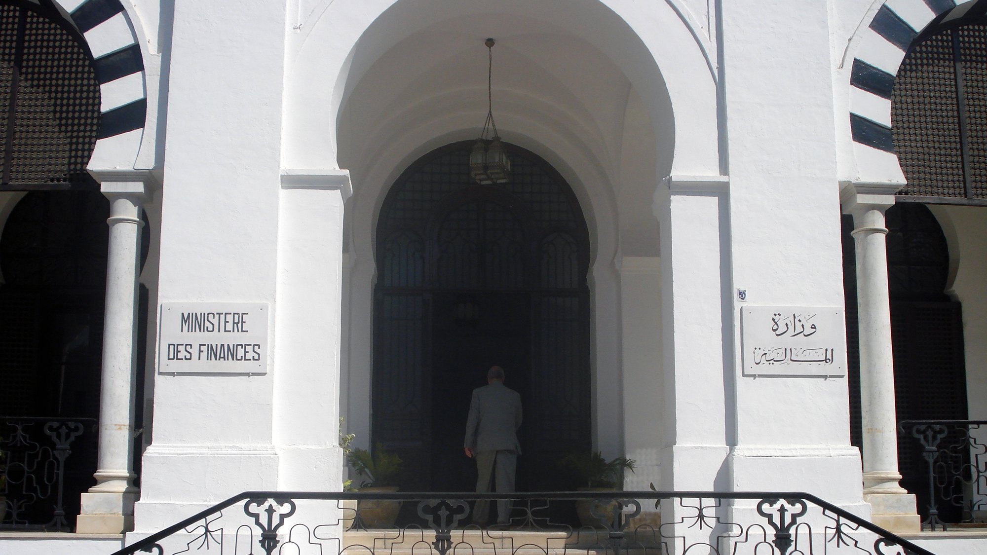 L'image montre l'entrée d'un bâtiment qui semble être le ministère des Finances, avec des inscriptions en français et en arabe. Le bâtiment est de style architectural classique, caractérisé par des murs blancs et des éléments décoratifs. On peut voir une grande porte d'entrée, entourée de fenêtres avec des grilles artistiques. À l'avant, une personne est visible, tournant le dos, s'approchant de l'entrée. Des plantes ornent l'espace autour des marches qui mènent à la porte.