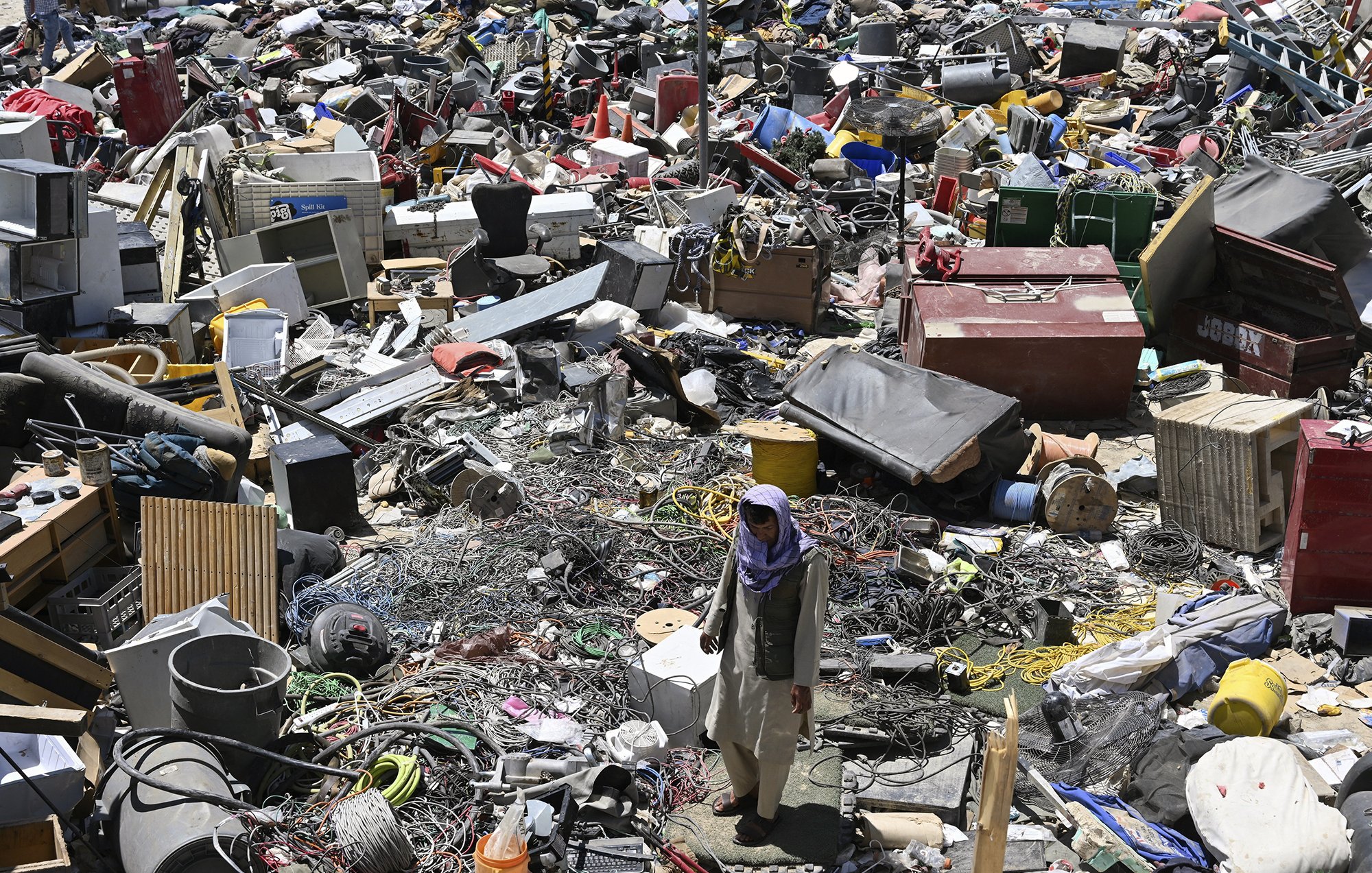 L'image montre un vaste amoncellement de déchets et de débris dans un espace encombré. On y voit des objets divers tels que des appareils électroménagers, des câbles, des pneus, et d'autres débris. Il y a aussi une personne qui marche au milieu de ce désordre, donnant une idée de l'échelle des détritus présents. L'environnement semble désordonné et négligé, illustrant une scène de récupération ou de désindustrialisation.