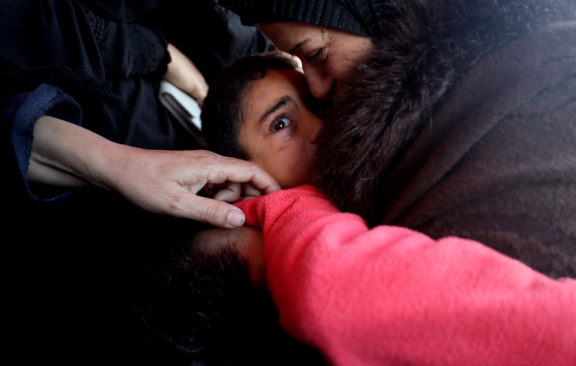 L'image montre un moment touchant entre un enfant et un adulte. L'adulte semble embrasser l'enfant, qui a l'air attentionné et gaze directement vers l'objectif. On peut aussi voir quelques mains, une d'elles touchant l'épaule de l'enfant. L'ambiance est empreinte d'émotion et de tendresse. Les vêtements des figures suggèrent un environnement austère.