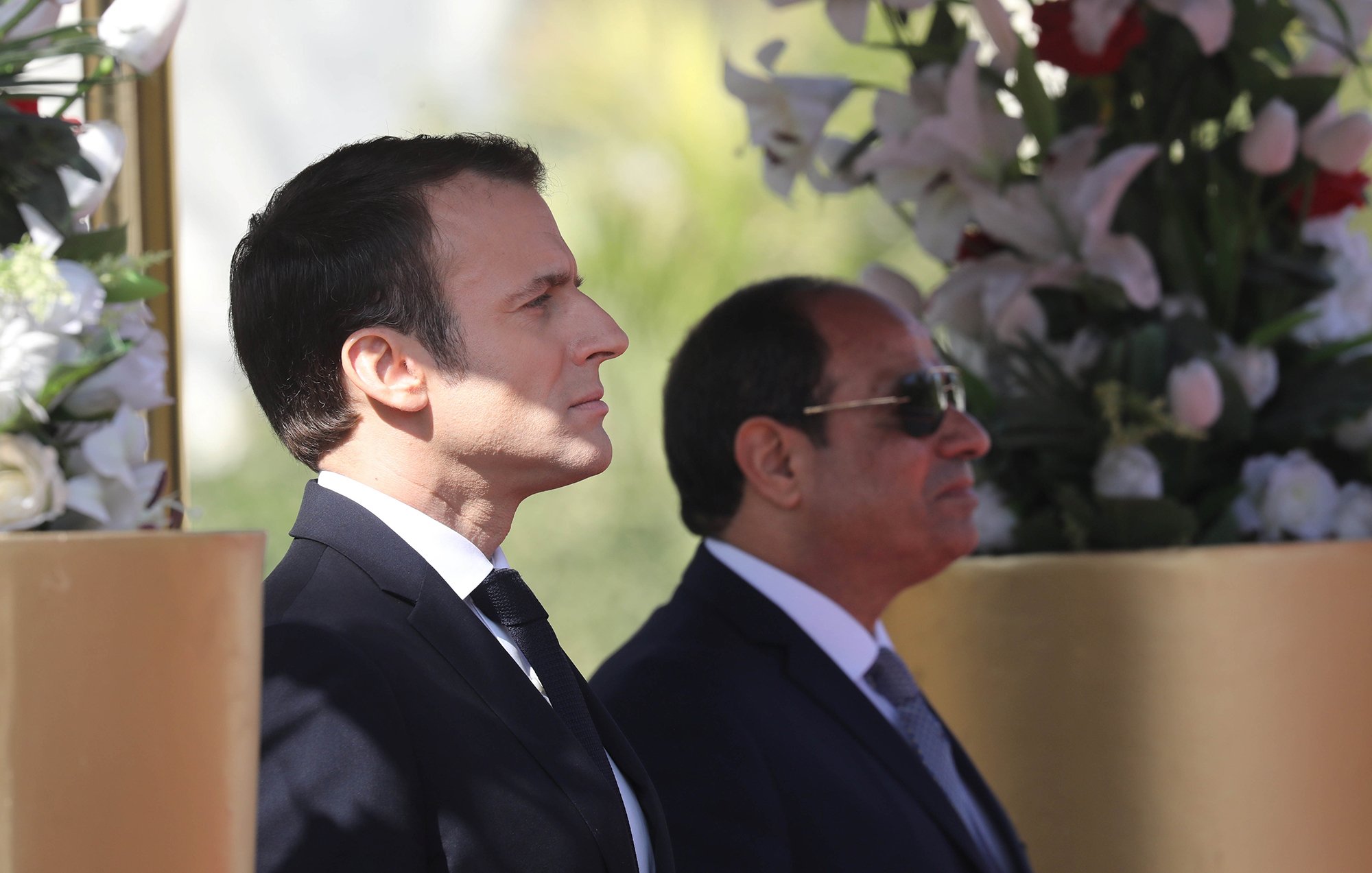 L'image montre deux hommes debout, apparemment lors d'un événement officiel. L'un d'eux est en costume sombre avec une cravate, il a une expression concentrée et regarde droit devant lui. L'autre homme, également en costume, porte des lunettes de soleil et semble observateur. Ils se tiennent près de décorations florales, suggérant un cadre cérémonieux.