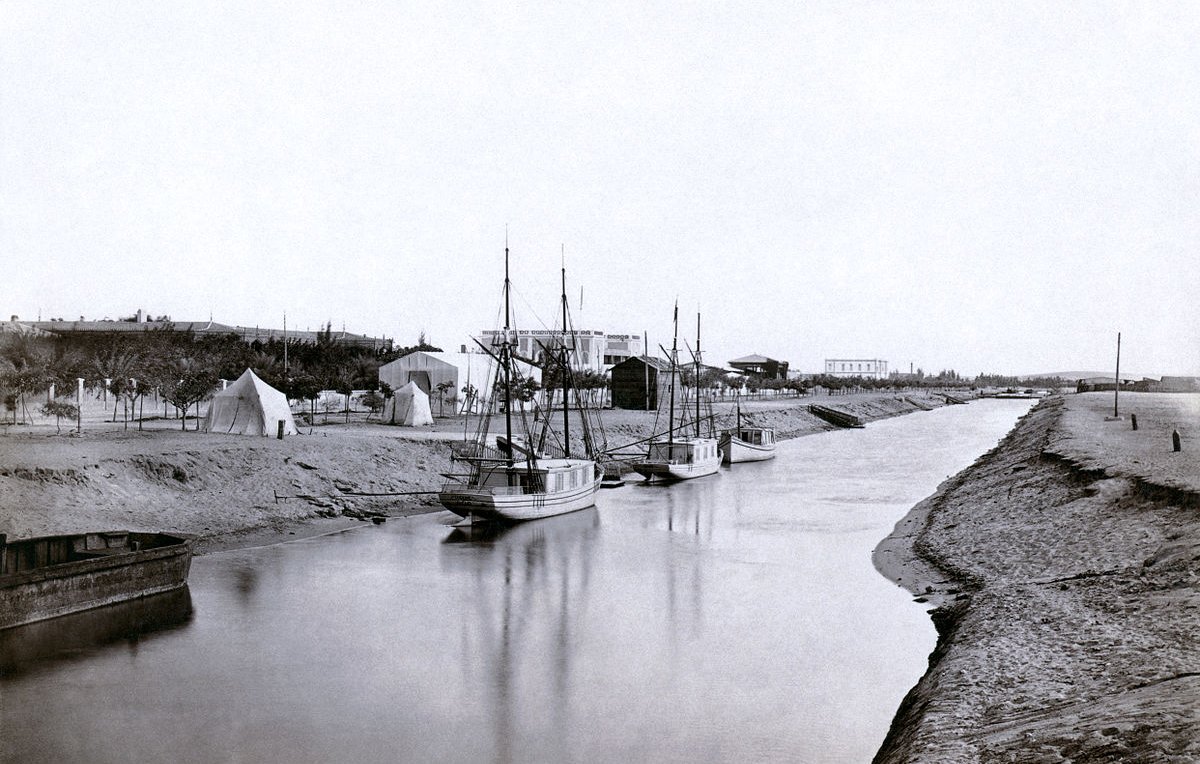 L'image montre une scène tranquille et nostalgique d'un canal ou d'une rivière. On y voit plusieurs bateaux amarrés, leur reflet se mêlant à l'eau calme. En arrière-plan, il y a des tentes et des structures qui évoquent une installation temporaire ou un camp. Le paysage environnant semble naturel, avec de la terre et des arbres. Cette photographie en noir et blanc donne une impression de sérénité et évoque une époque passée.