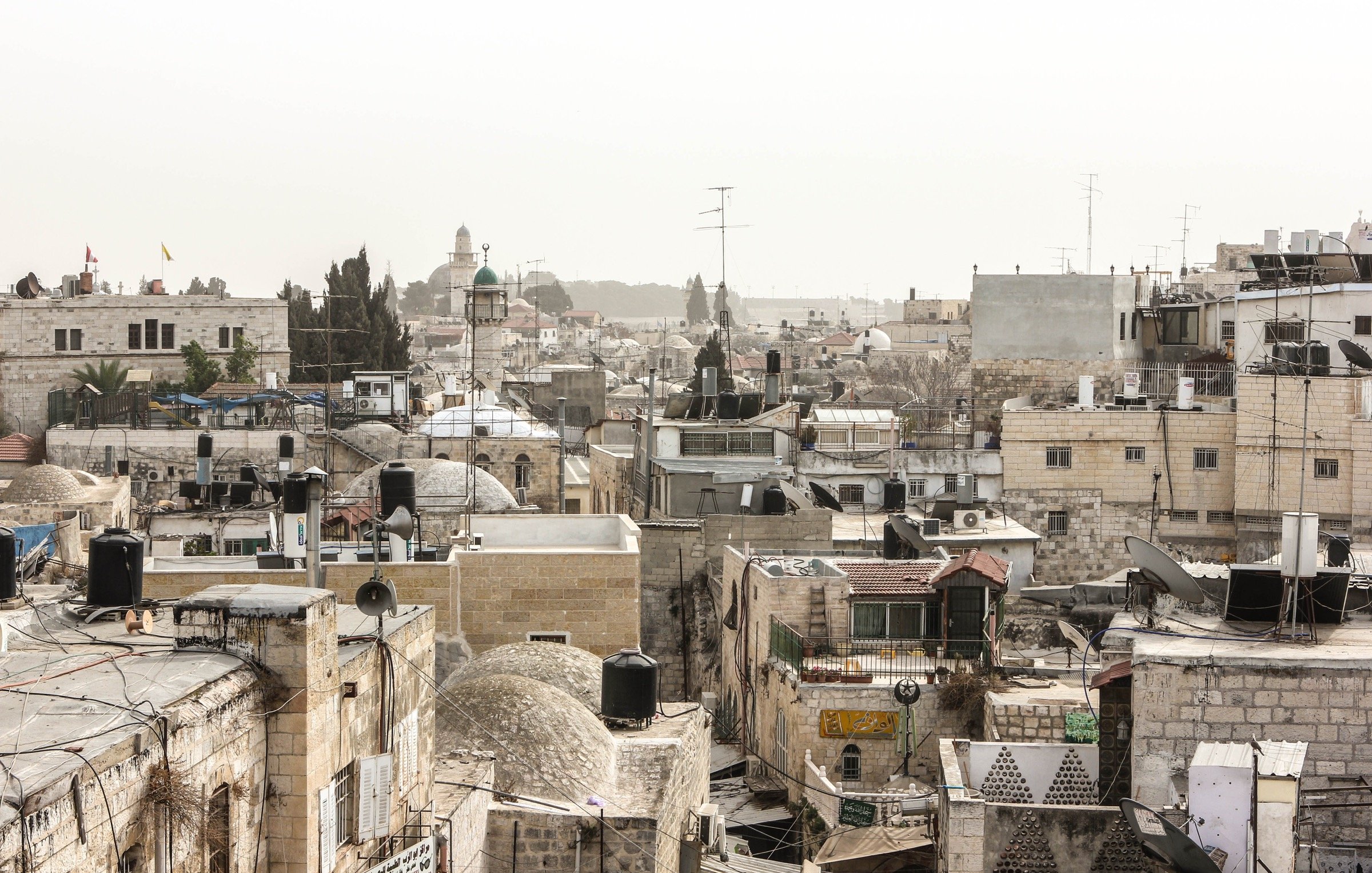 L'image montre un paysage urbain d'une ville ancienne, probablement Jérusalem. On peut voir une multitude de bâtiments en pierre avec des toits en tôle et des dômes, créant une texture variée. Les structures sont serrées les unes contre les autres, et quelques antennes de télévision sont visibles. Au loin, se distingue un minaret ou une coupole, indiquant la présence d'une mosquée. L'atmosphère semble un peu brumeuse, donnant une impression de chaleur et d'ancienneté à la scène.