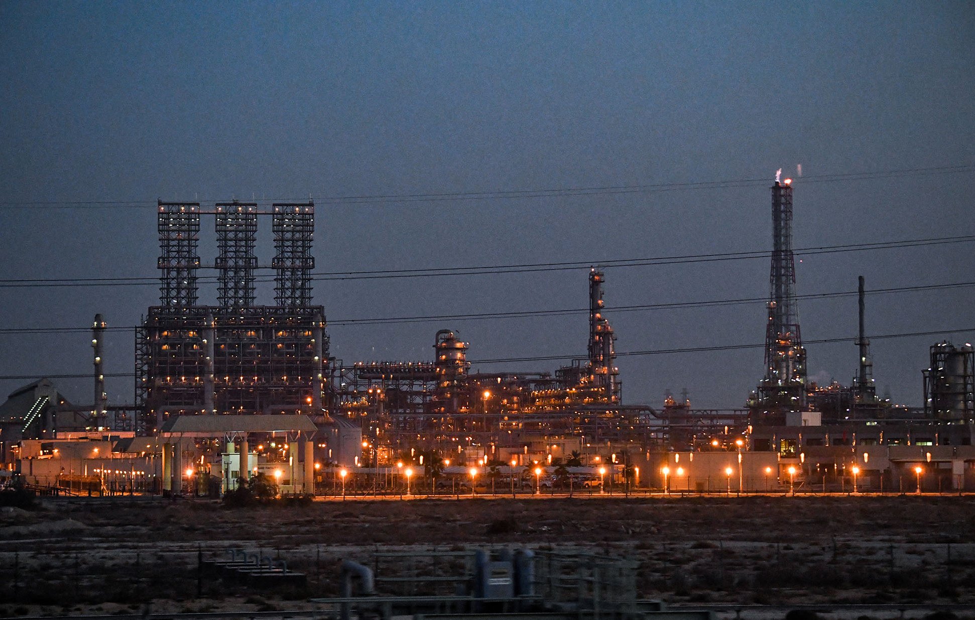L'image montre une grande installation industrielle, probablement une raffinerie ou une usine de traitement, éclairée par de nombreuses lumières à la tombée de la nuit. On peut voir de hautes structures vertes et des cheminées qui s'élèvent dans le ciel. L'environnement autour semble désertique, avec peu de végétation visible. Les lignes électriques traversent le premier plan, ajoutant à l'atmosphère de l'image. Les lumières orange et blanches des installations créent un contraste avec le ciel sombre.