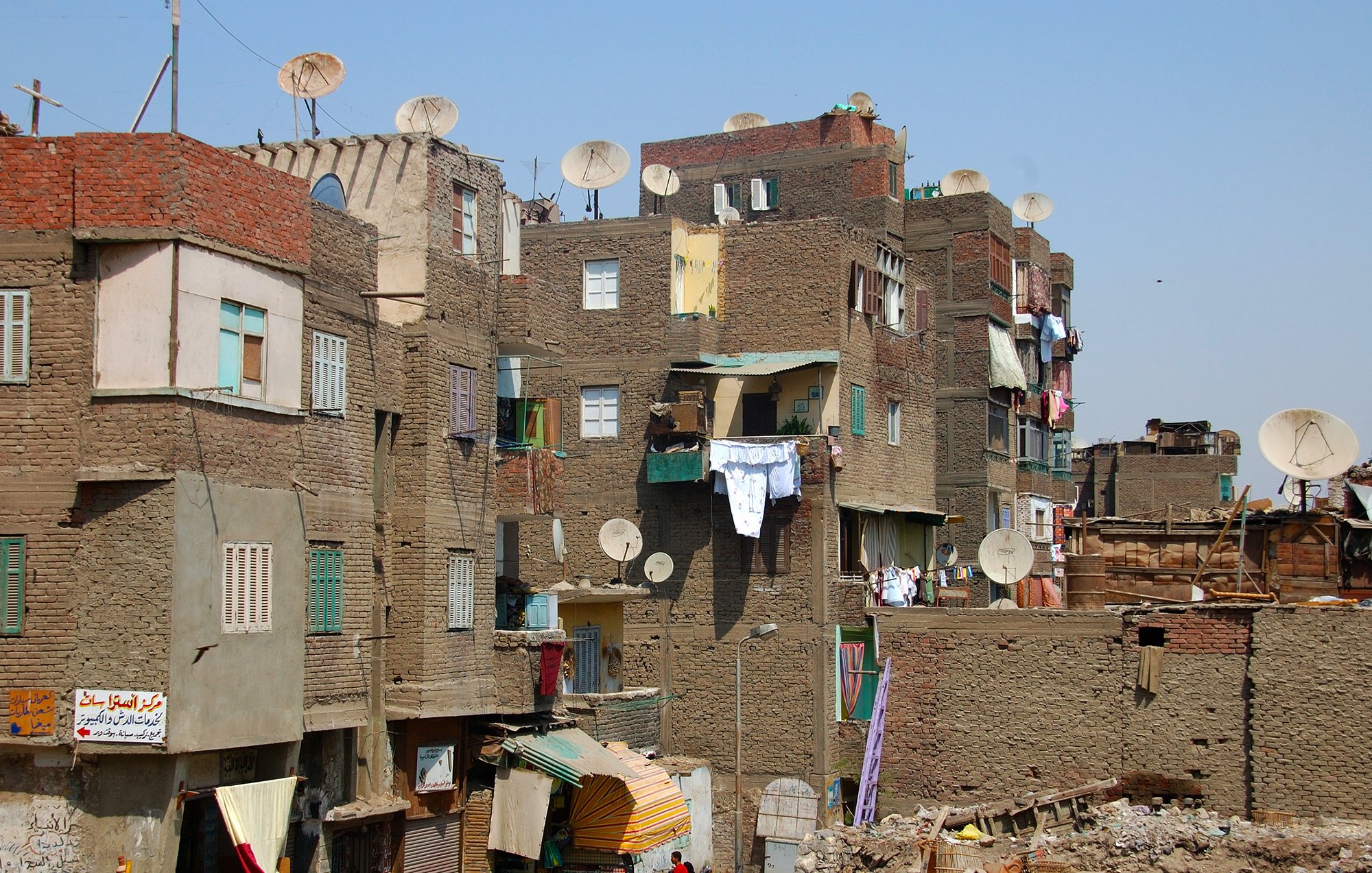 L'image montre un paysage urbain d'un quartier populaire. On peut voir des bâtiments en briques de terre, souvent de plusieurs étages, avec des façades colorées. De nombreuses antennes parabolique sont installées sur les toits. Les vêtements sont accrochés à l'extérieur pour sécher. L'atmosphère semble animée, avec des signes de vie quotidienne. Le ciel est dégagé, ce qui donne une impression de chaleur.