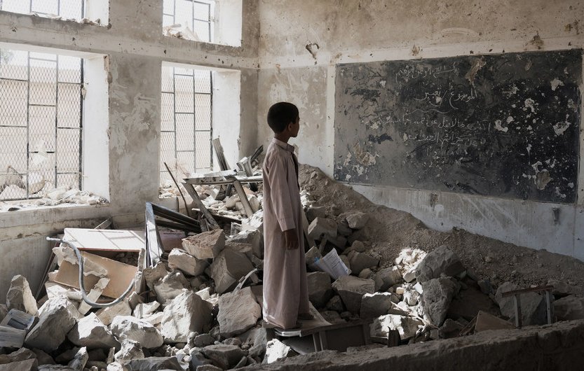 L'image montre un enfant se tenant dans une salle de classe en ruines. Le sol est jonché de débris, tandis que des morceaux de murs effondrés et des meubles renversés témoignent de la destruction. Un tableau noir, partiellement visible, porte des inscriptions, bien que l'environnement soit dégradé. La lumière pénètre à travers les fenêtres, créant un contraste avec l'atmosphère sombre et désolée de la pièce. Cette scène évoque une profonde sensation de perte et de désespoir.