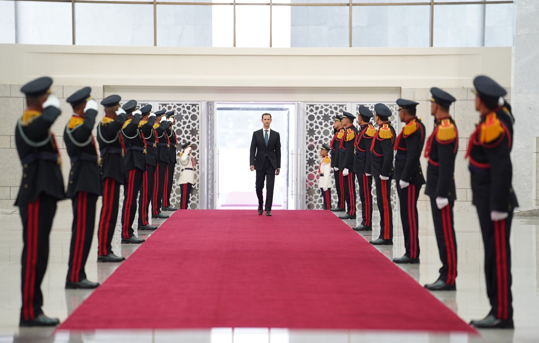 L'image montre une scène formelle dans un hall décoré. Au centre, un homme en costume marche sur un tapis rouge, flanqué de gardes en uniforme qui lui rendent les honneurs en levant la main au-dessus du front. Les gardes sont en rangées de chaque côté, portant des uniformes noirs avec des accents colorés. L'arrière-plan présente des murs en marbre et une grande ouverture lumineuse. L'atmosphère est solennelle et cérémonielle.