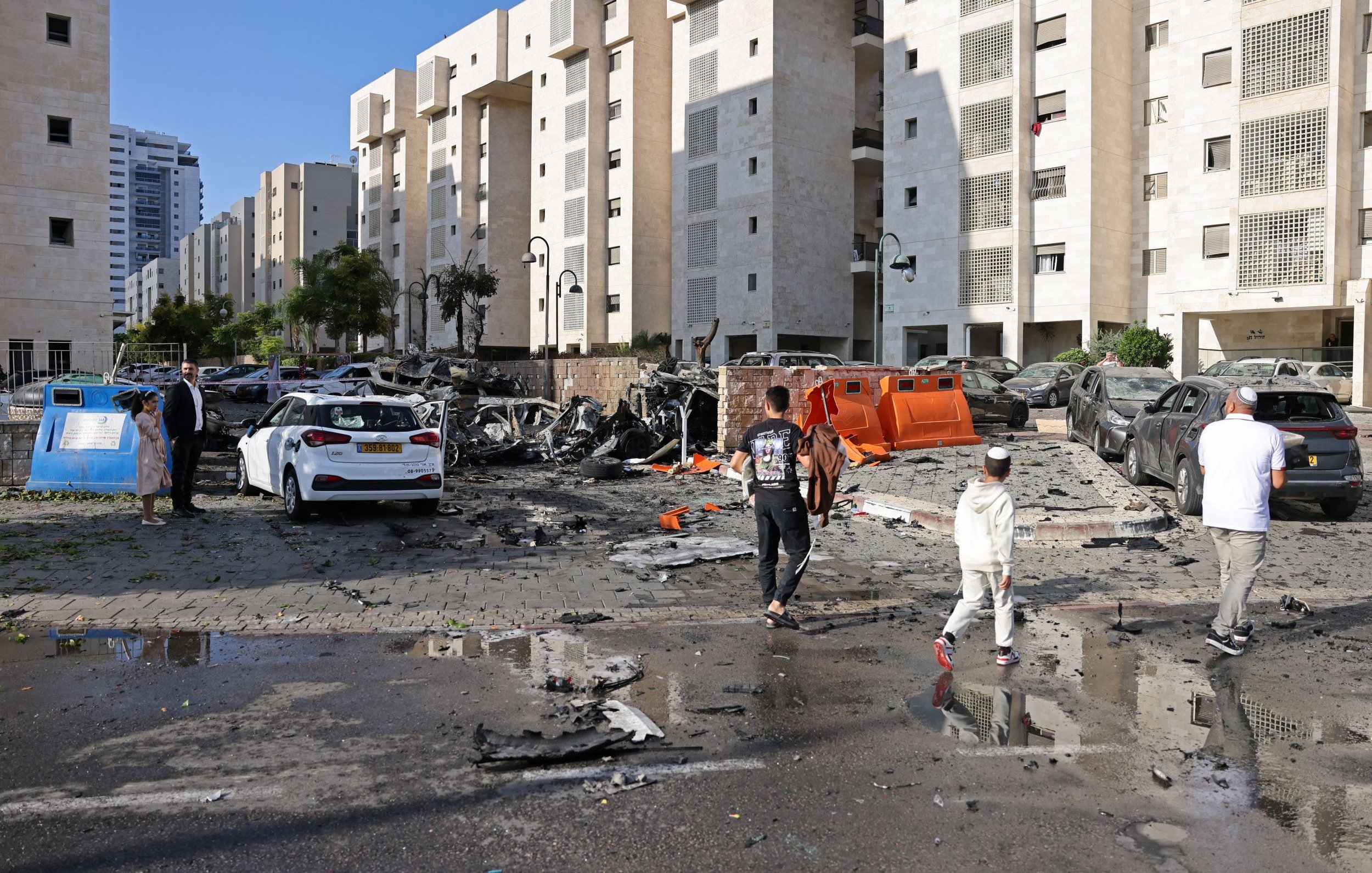 L'image montre une scène de destruction urbaine. On peut voir plusieurs véhicules endommagés, certains complètement détruits. Le sol est couvert de débris, et il semble qu'une explosion ou un événement violent ait eu lieu. En arrière-plan, il y a des bâtiments modernes. Des personnes marchent sur la route, apparemment en train d'évaluer les dégâts. Le ciel est clair, ce qui contraste avec l'image de désolation.