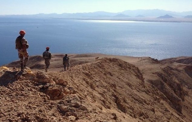 L'image montre un paysage désertique avec des militaires en patrouille sur une crête surplombant une étendue d'eau. Le terrain est aride et rocheux, avec des montagnes en arrière-plan. Les soldats portent des uniformes et des casques, et semblent observer l'horizon, avec un ciel clair et ensoleillé qui ajoute à l'atmosphère de la scène.