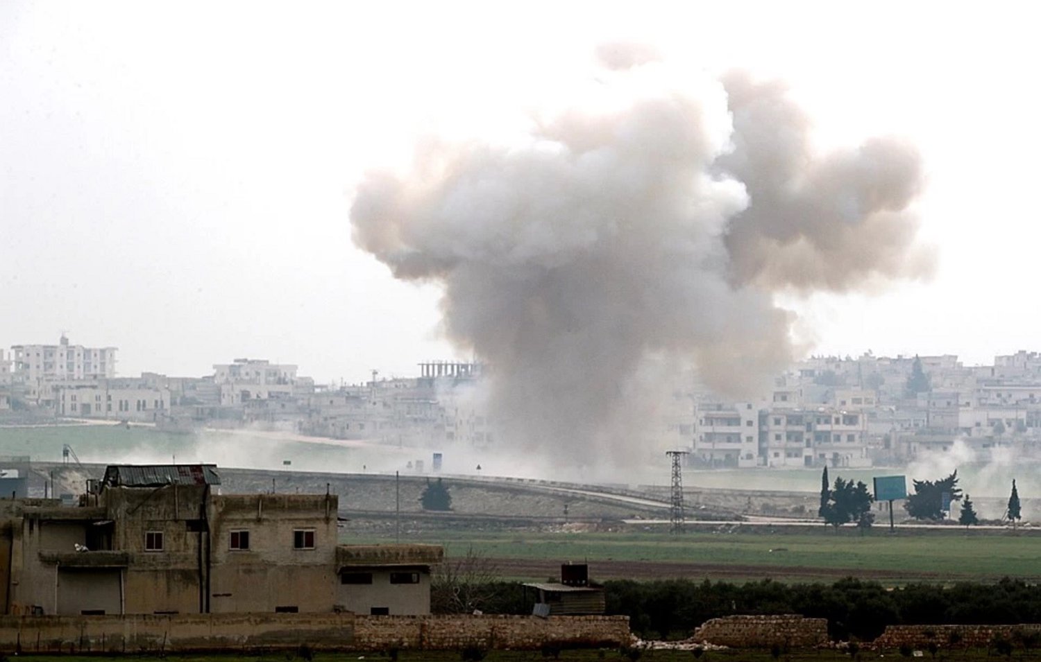 L'image montre un paysage de guerre, avec une explosion qui génère un nuage de fumée sombre s'élevant dans le ciel. Au premier plan, on peut apercevoir des bâtiments endommagés, tandis qu'à l'arrière-plan, une ville est visible, probablement touchée par le conflit. L'atmosphère dégage une impression de destruction et de chaos, illustrant les conséquences d'une situation de conflit armé.