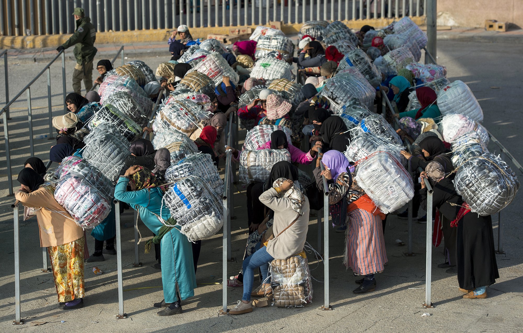 L'image montre un groupe de personnes, principalement des femmes, qui portent de grands fardeaux enveloppés dans des filets ou des tissus. Elles se trouvent dans un environnement urbain, probablement près d'un point de passage ou d'une frontière. La plupart des femmes sont vêtues de vêtements traditionnels et portent des foulards. Elles semblent attendre ou se regrouper, tandis qu'en arrière-plan, on aperçoit un soldat ou un garde. Cette scène évoque des thèmes de travail, de migration ou de transport de marchandises.