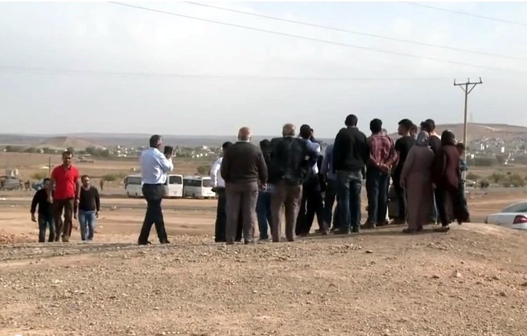 L'image montre un groupe de personnes rassemblées sur un terrain semi-aride. Ils semblent observer quelque chose au loin, peut-être un événement ou une scène particulière. L'arrière-plan est dégagé, avec des collines et des bâtiments visibles au loin. Il y a des véhicules stationnés, dont une voiture à droite. L'atmosphère suggère une interaction entre les gens, qui semblent concentrés sur ce qu'ils regardent. Des câbles électriques sont visibles, ajoutant à l'ambiance rurale de la scène.