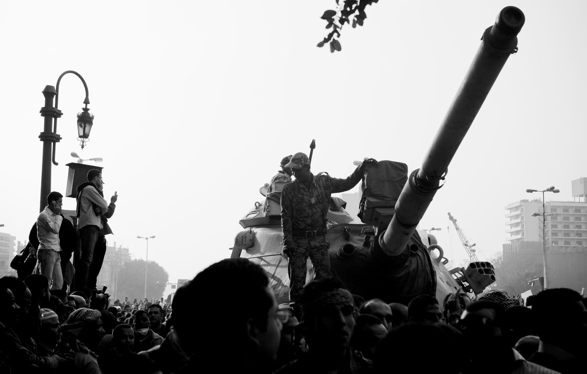 L'image montre une scène de manifestation avec un tank au premier plan. Des soldats sont visibles, dont un se tient debout sur le tank, levant un bras en signe de protestation ou de triomphe. Au premier plan, on aperçoit une foule de manifestants, certains prenant des photos ou filmant la scène. L'atmosphère est tendue, peut-être en raison de la situation politique. L'image est présentée en noir et blanc, ce qui renforce le côté dramatique de la scène.