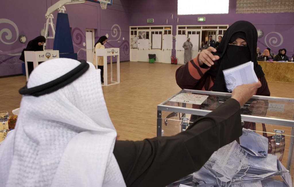 L'image montre une scène d'élection dans un environnement intérieur. Au premier plan, un homme portant un kandora et un ghutrah se tient derrière une urne électorale. Il regarde une femme voilée qui tend un bulletin de vote, le visage partiellement caché par un niqab. En arrière-plan, on peut voir d'autres personnes et des isoloirs, indiquant que le processus de vote est en cours dans cet espace, qui semble être une salle de sport ou un centre communautaire. La décoration du mur est colorée, ajoutant une atmosphère dynamique à l'événement.